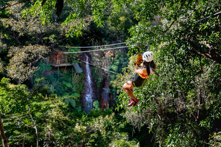 Person zip-lining through forest with waterfall in background, wearing a helmet and safety gear.