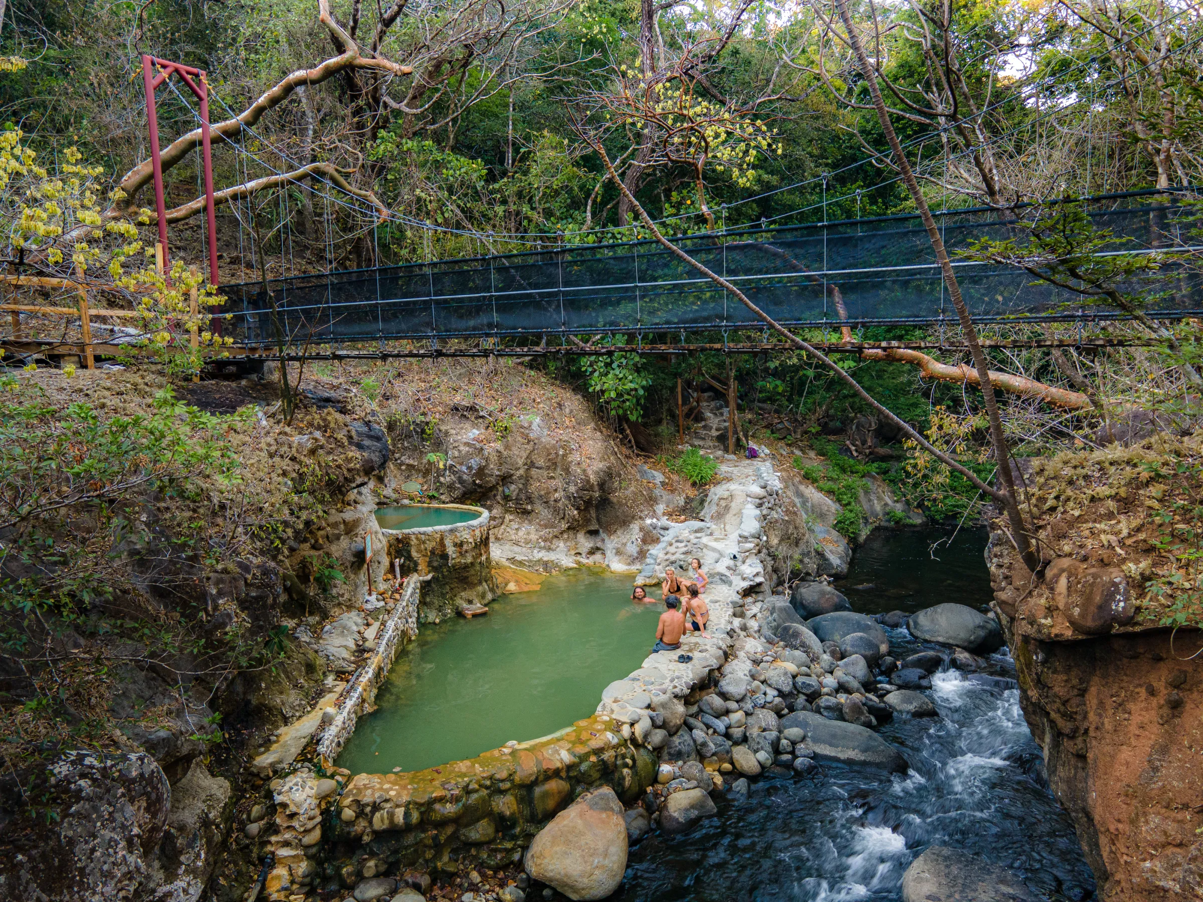 People relax in hot springs below a footbridge in a lush, rocky landscape.