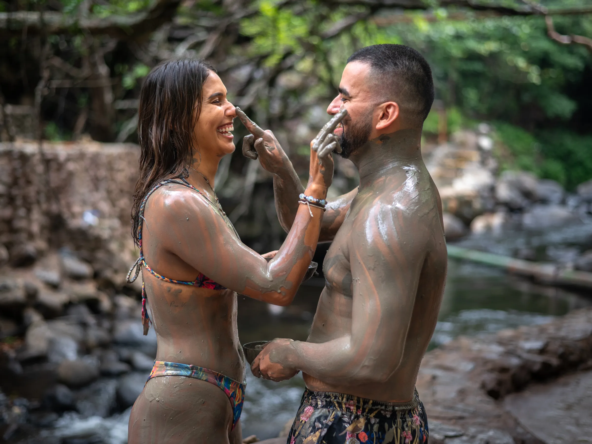 Two people covered in mud smile and apply mud on each other's faces near a river.