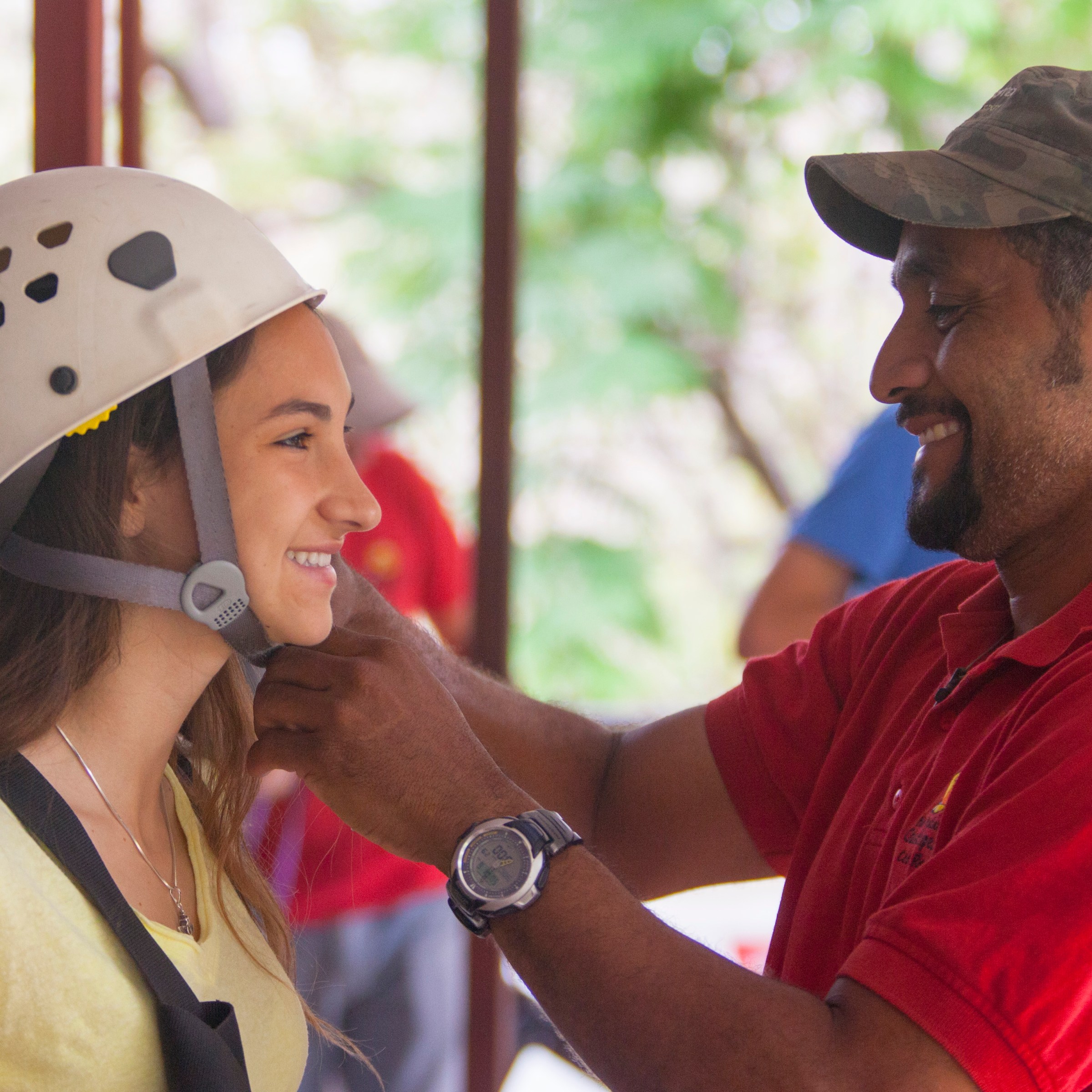 Man in red shirt helps woman adjust helmet strap outdoors.