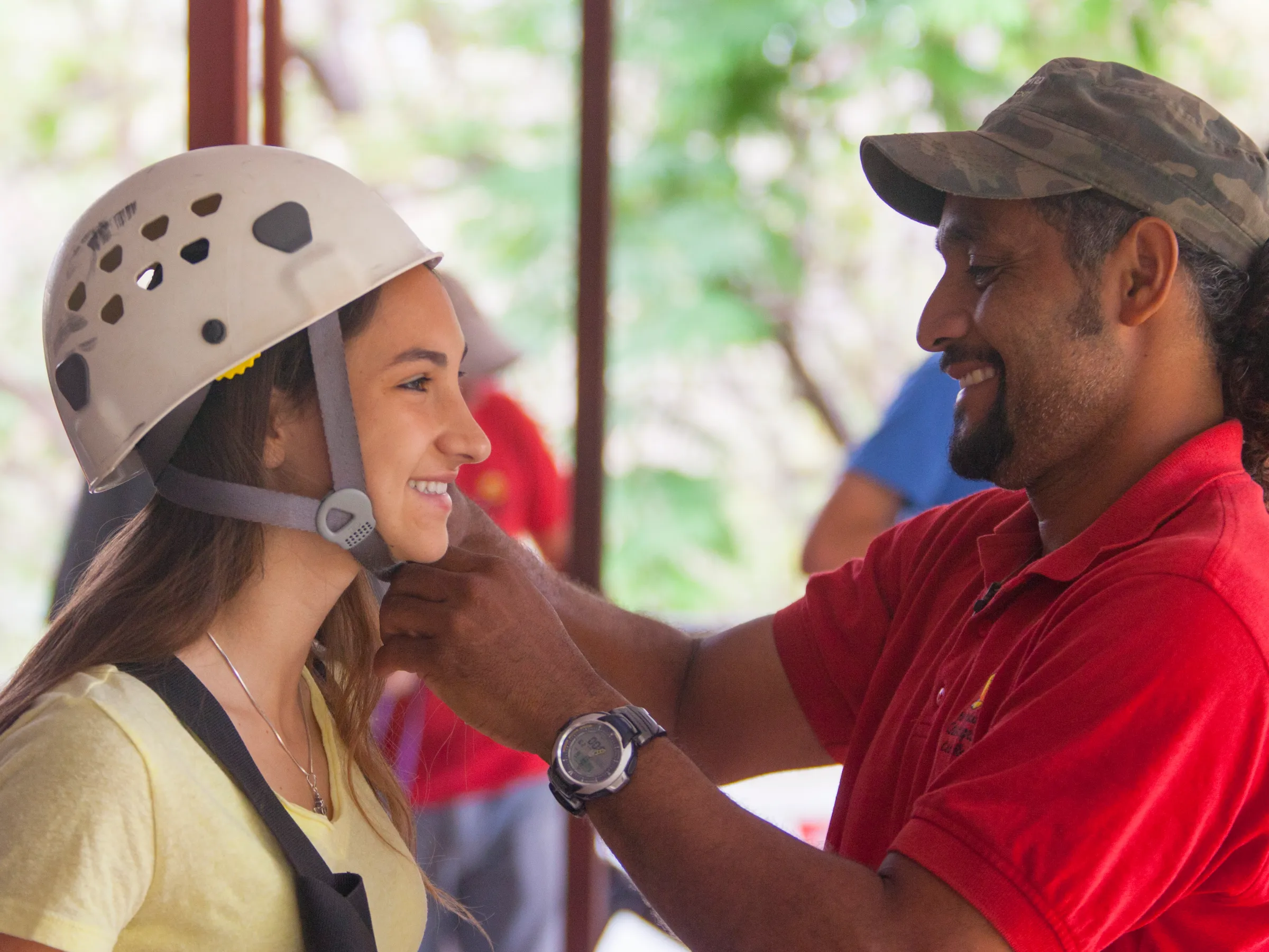 Man in red shirt helps woman adjust helmet strap outdoors.