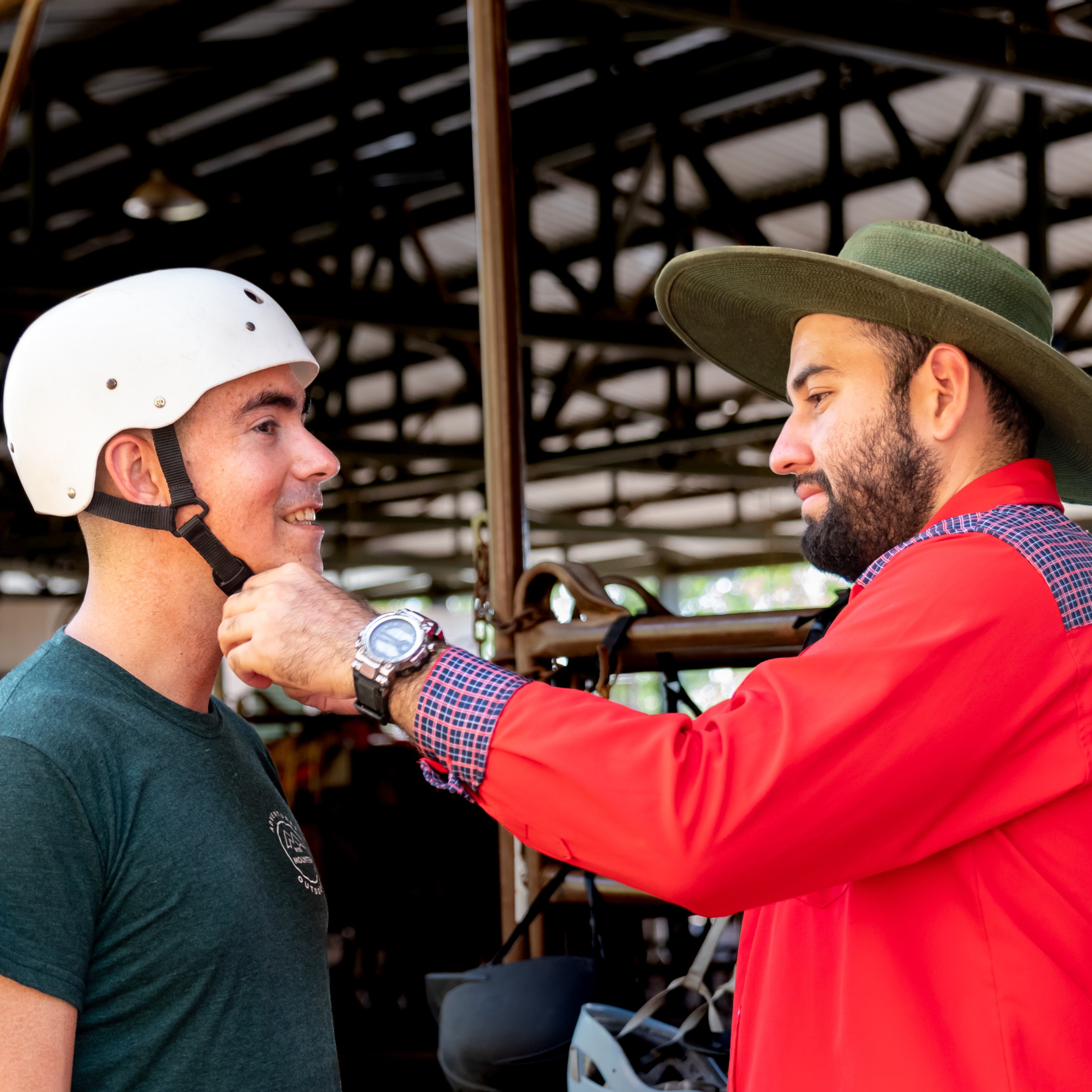 Man in hat helps another man adjust a white helmet in a barn setting.