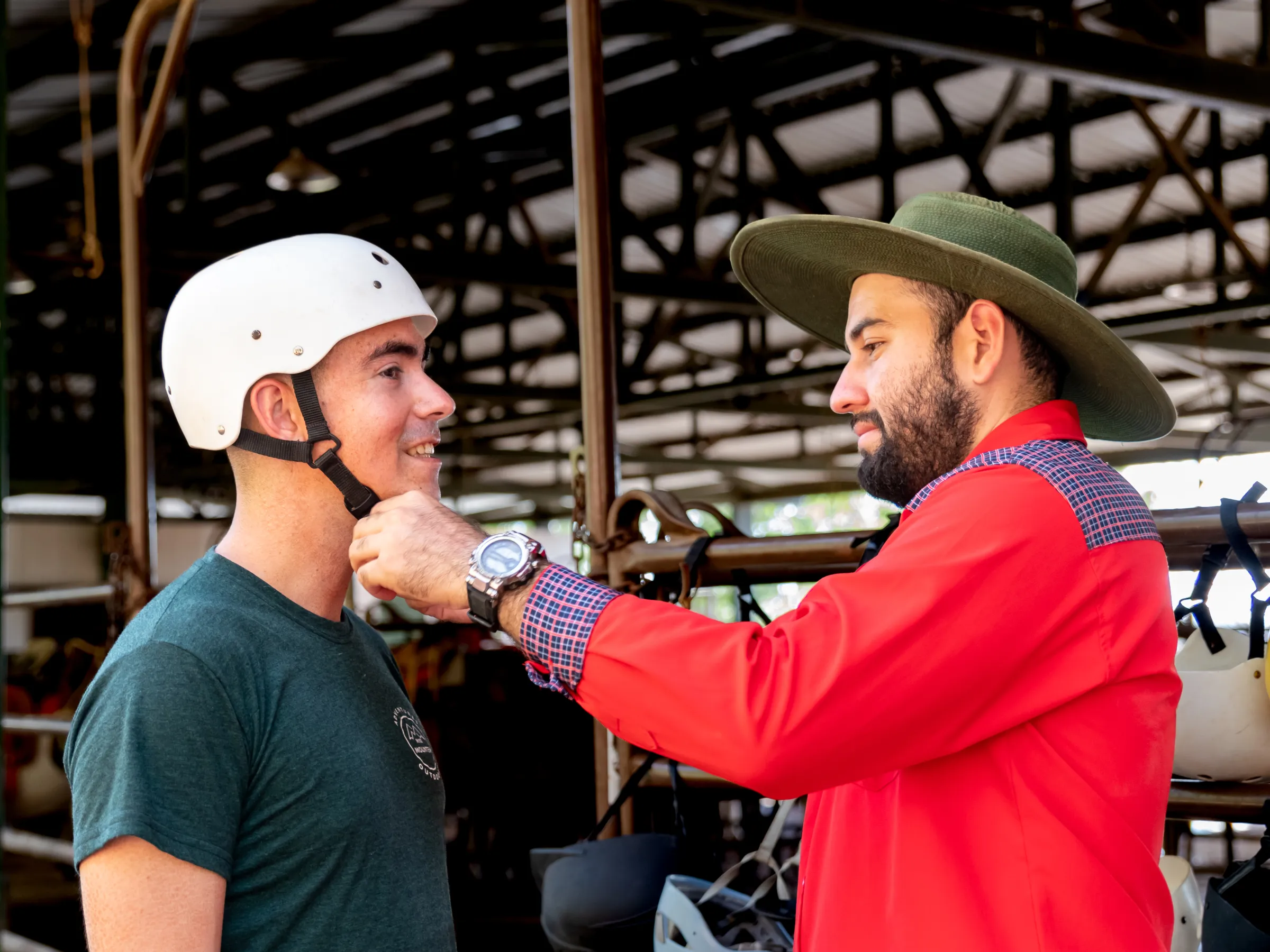 Man in hat helps another man adjust a white helmet in a barn setting.