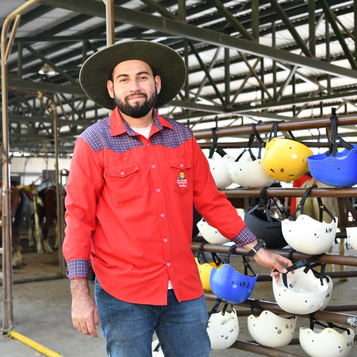 Man in red shirt and hat stands by helmets in an equestrian stable.
