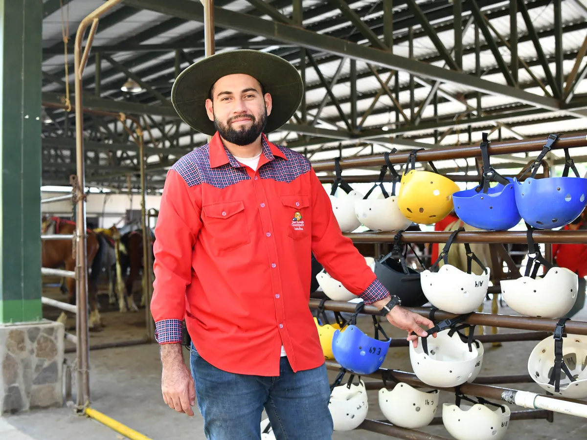 Man in red shirt and hat stands by helmets in an equestrian stable.