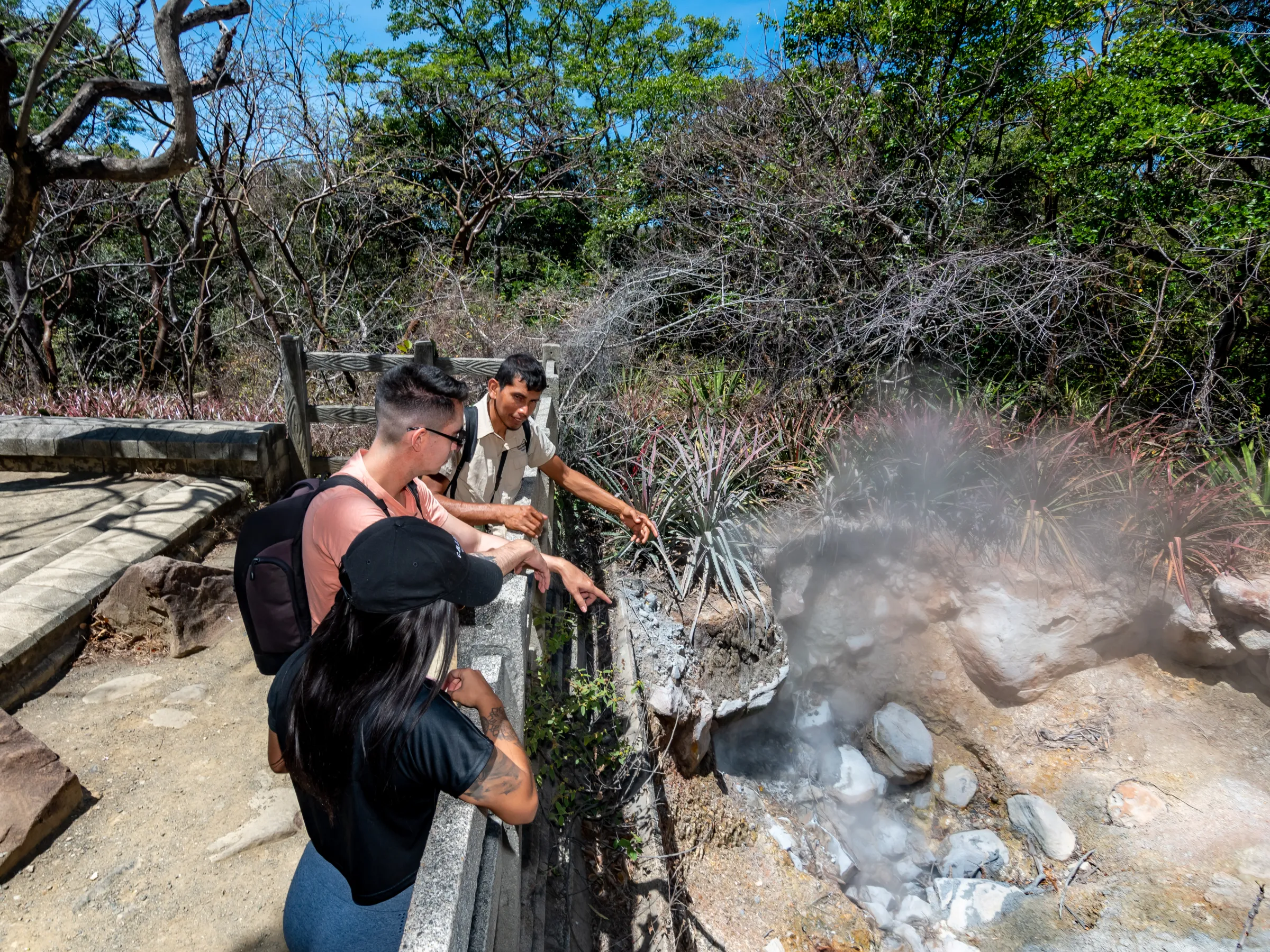 Three people observing steam rising from a geothermal area in a forest.