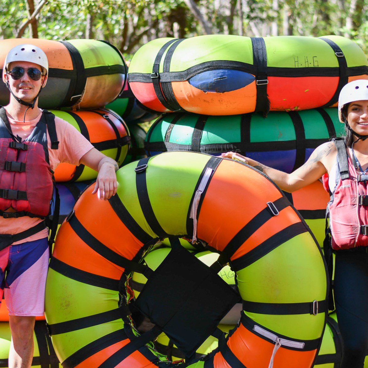 Two people in helmets and life vests stand with colorful inflatable tubes outdoors.