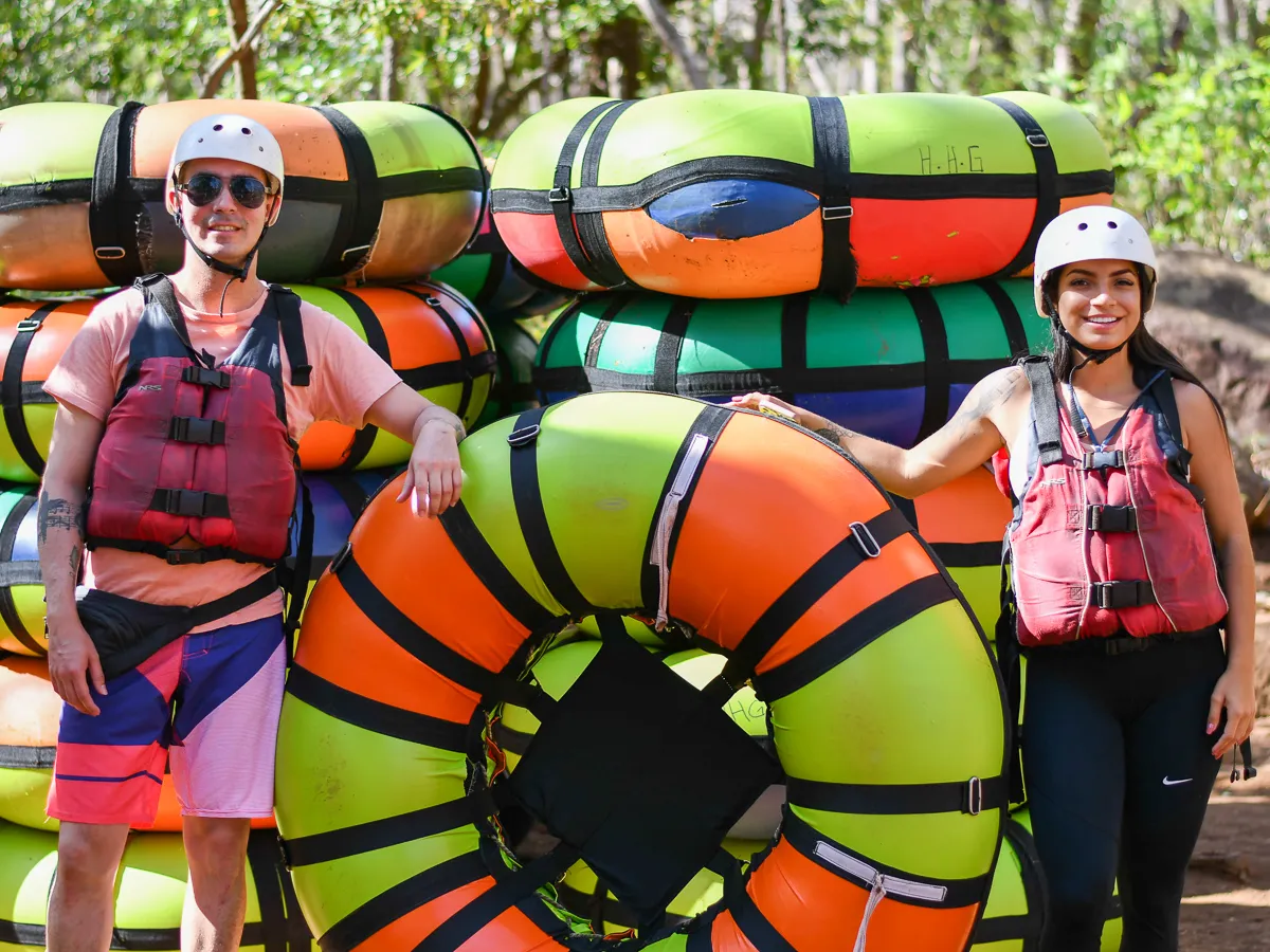 Two people in helmets and life vests stand with colorful inflatable tubes outdoors.