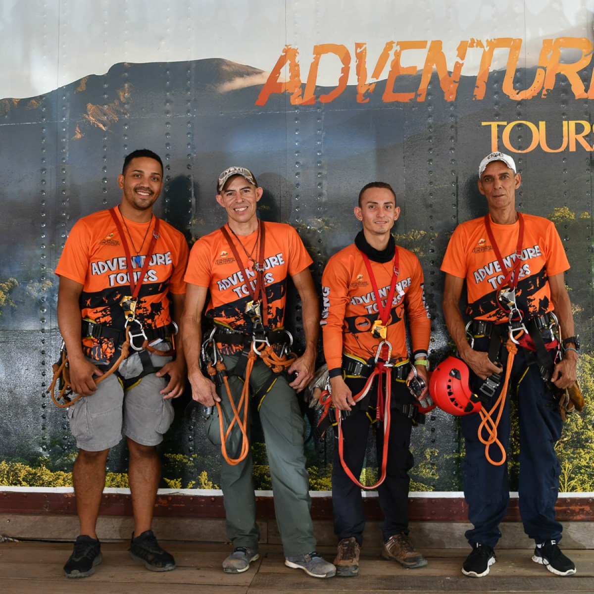 Four men in orange adventure gear posing in front of an 'Adventure Tours' sign.