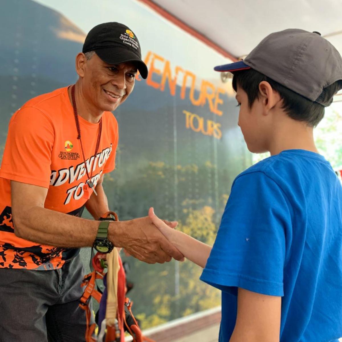 Man in orange shirt shakes hands with a boy in a blue shirt and cap.