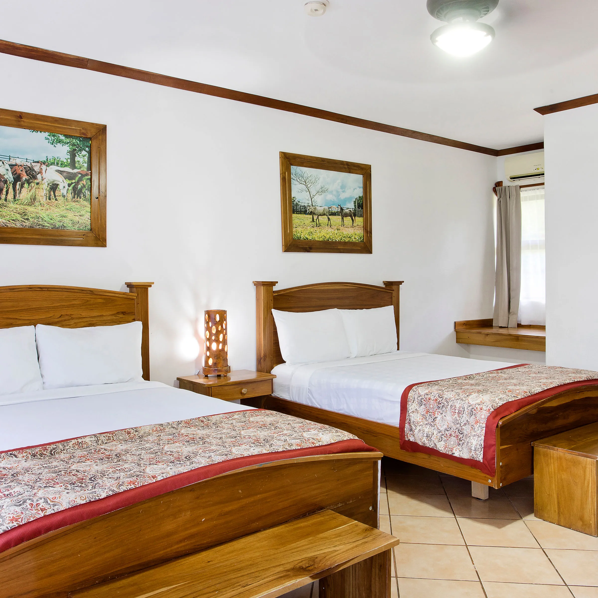 Hotel room with two wooden beds, decorative bedcovers, paintings, and a small side table with a lamp.