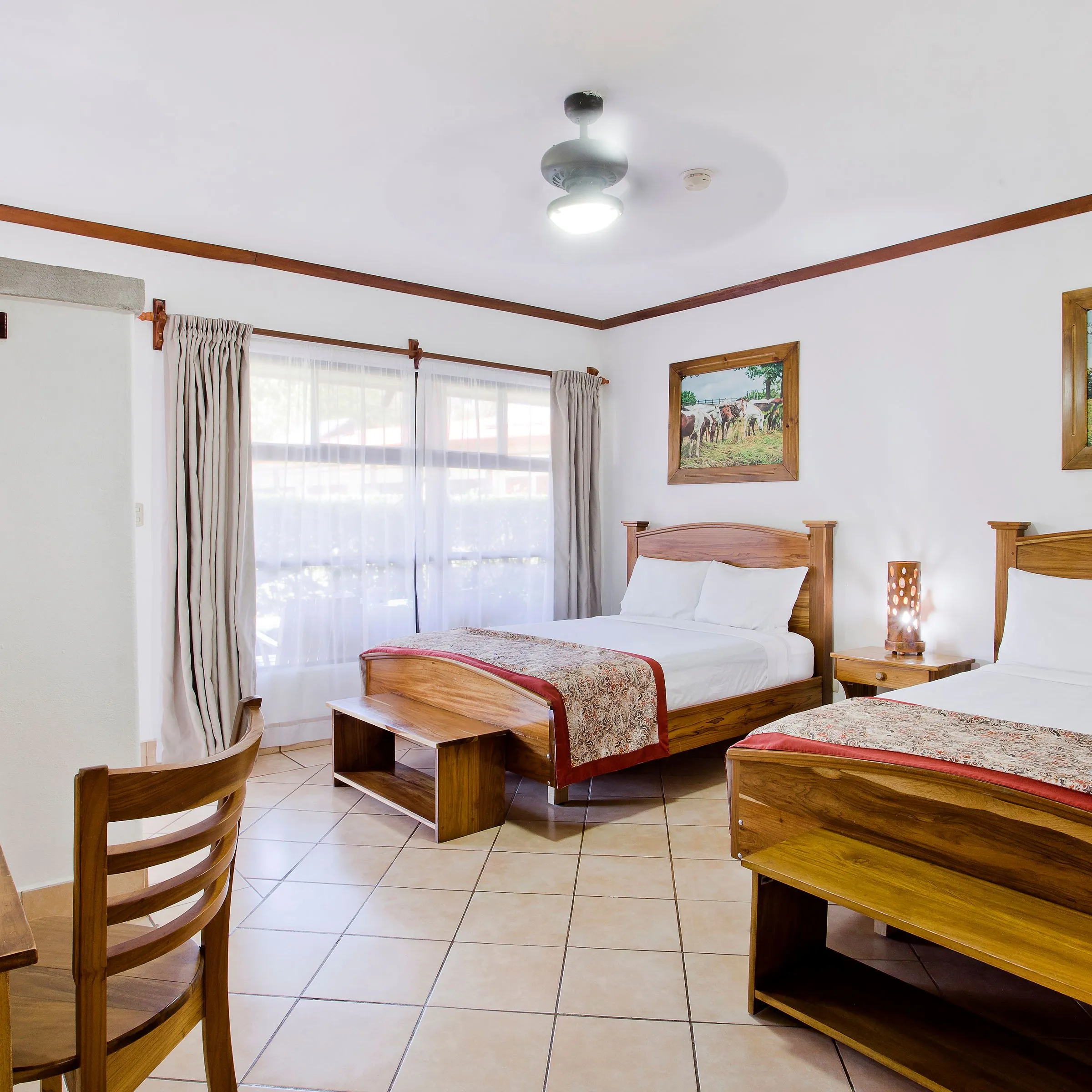 Hotel room with two beds, wooden furniture, paintings, a window with curtains, and ceiling fan.