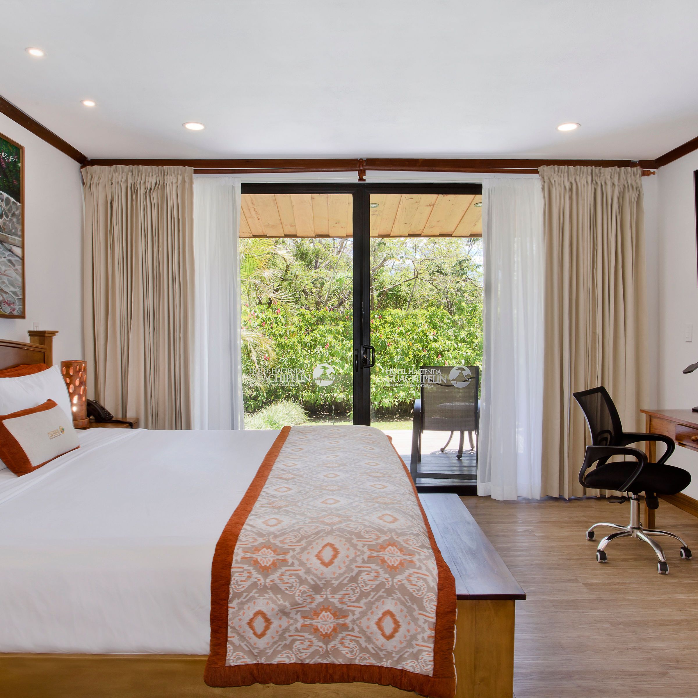 Bright hotel room with a large bed, desk, chair, TV, painting, and glass doors opening to a patio.
