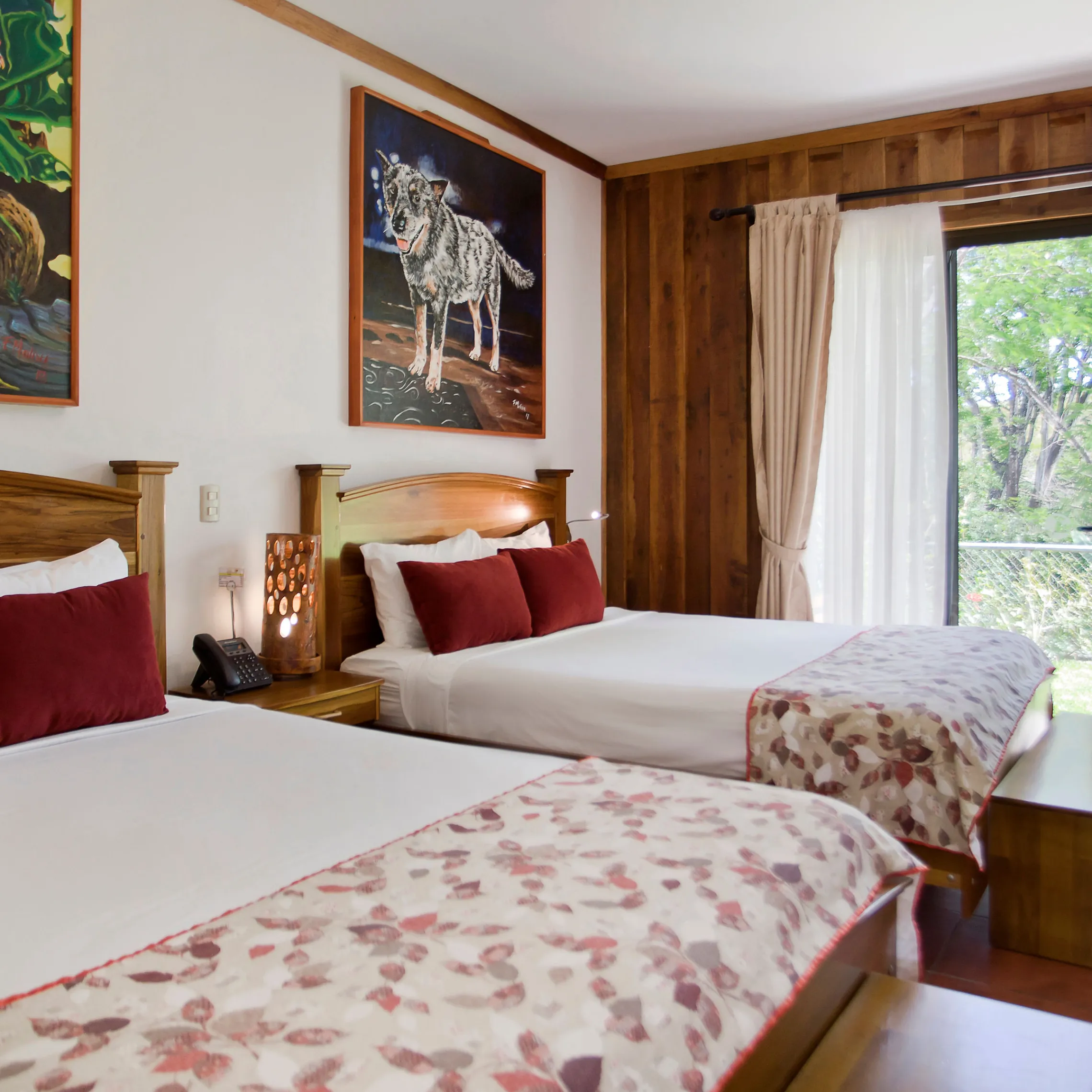 Room with two beds, paintings of a rooster and a wolf, and large window overlooking greenery.