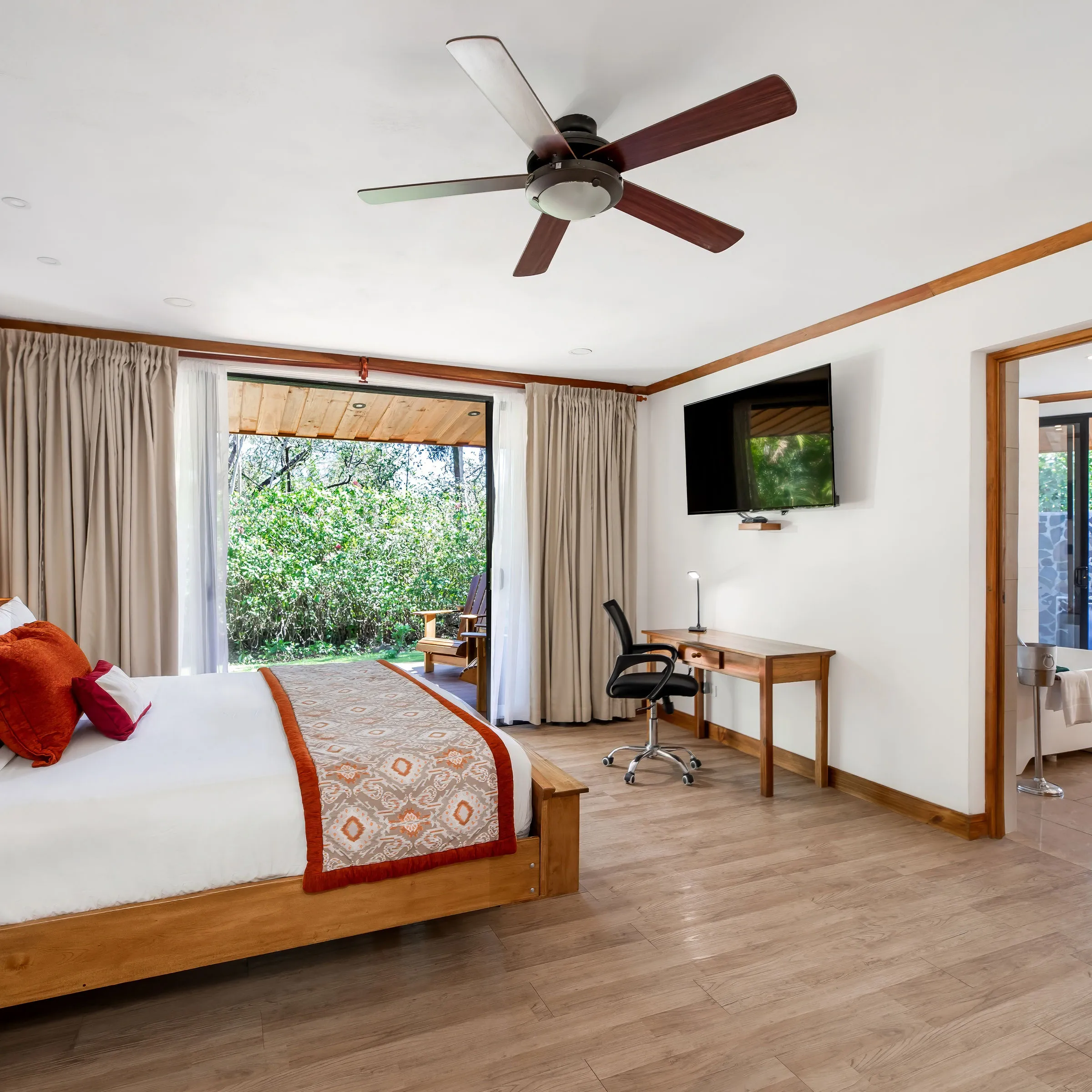 Modern bedroom with a large bed, ceiling fan, desk, TV, and bathroom with stone walls and outdoor view.