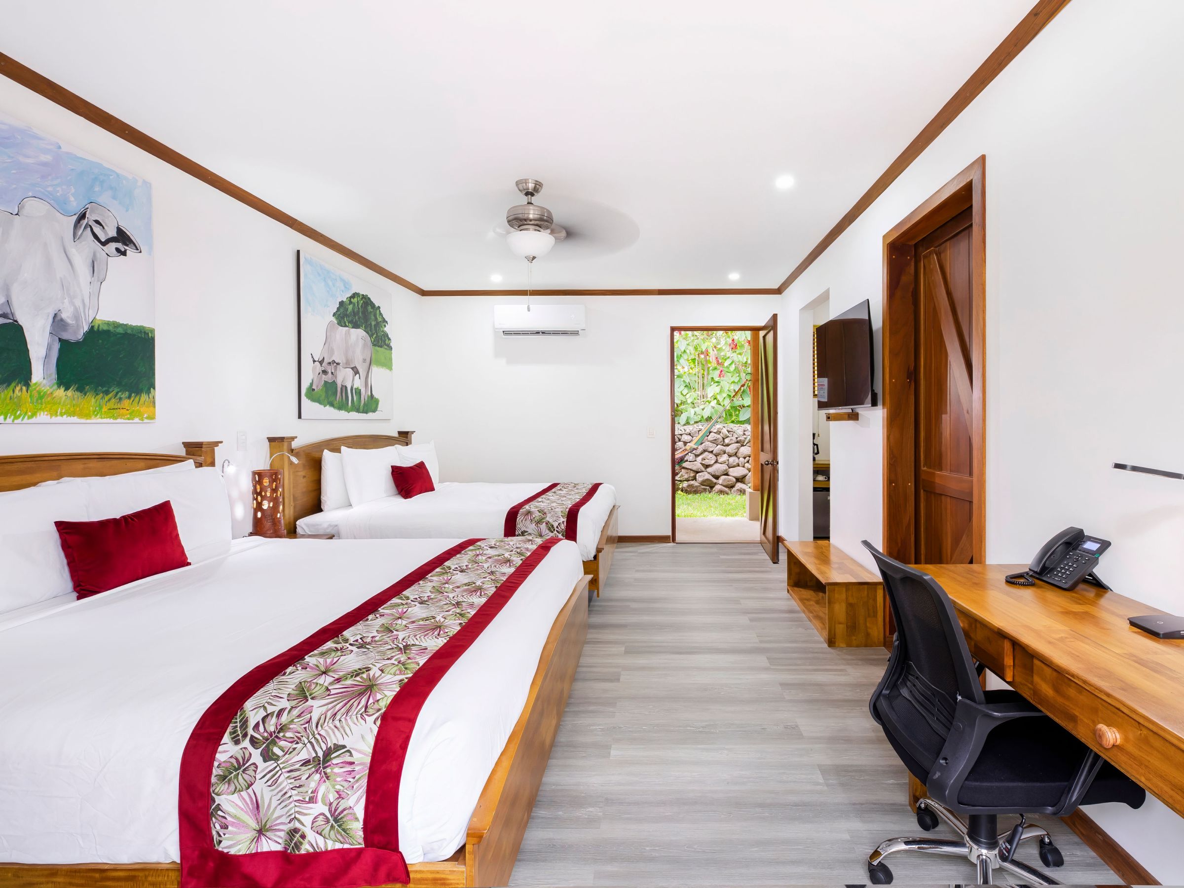 Hotel room with two beds, cow paintings, wood furniture, and a desk, facing door to garden.