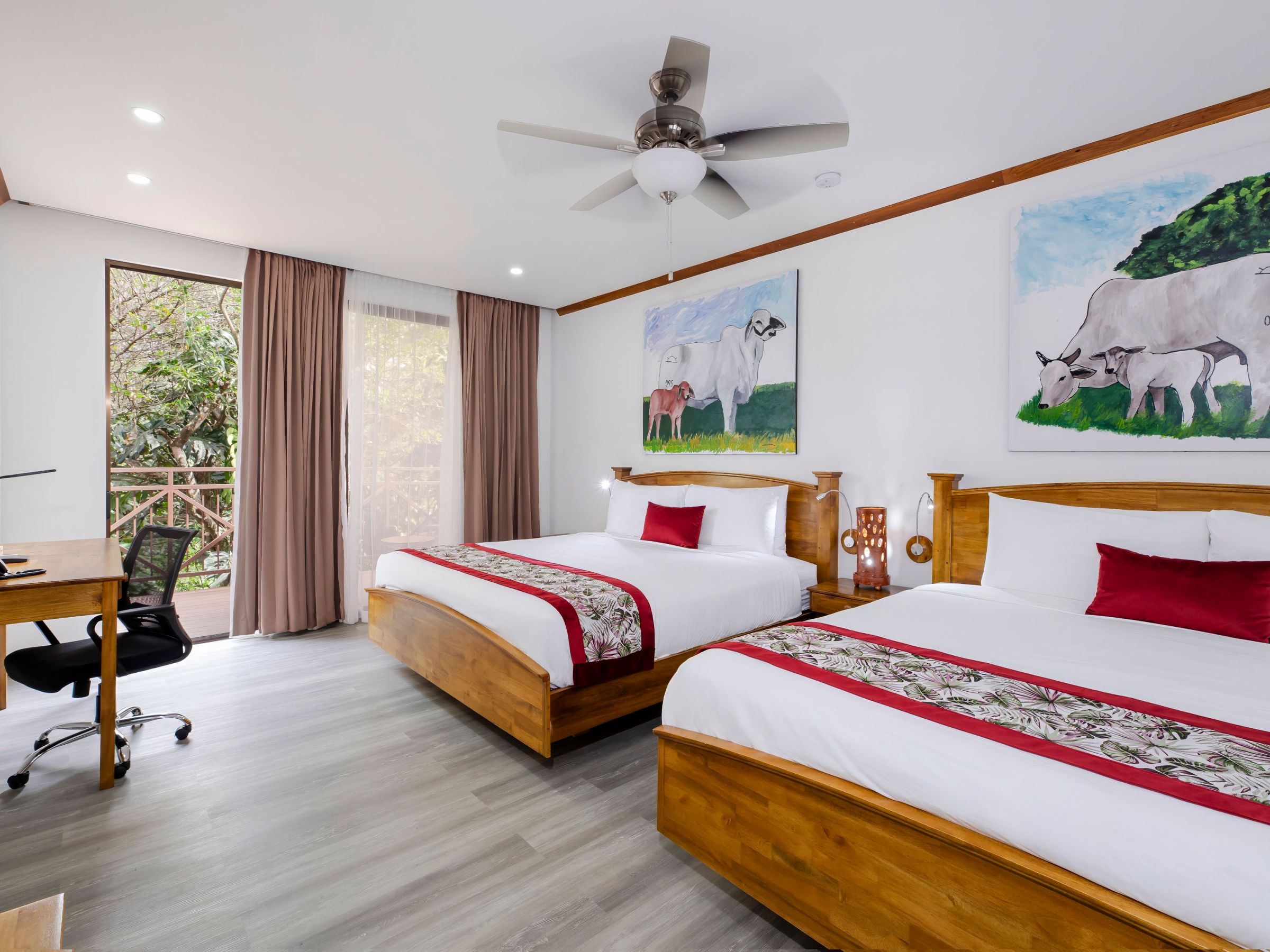 Room with two beds, paintings of cows, desk, chair, fan, and balcony view with curtains.