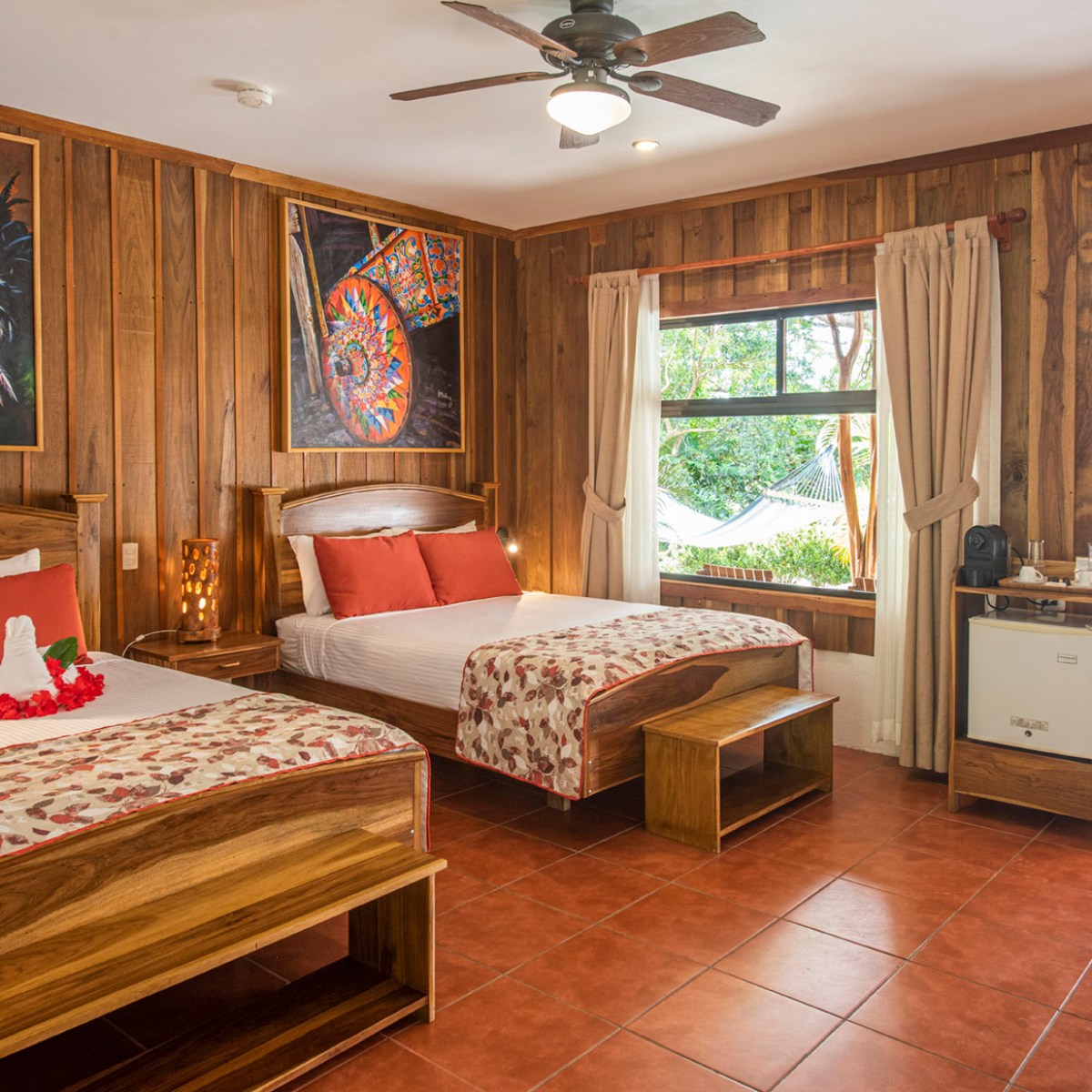 Wooden hotel room with two beds, rooster artwork, and garden view through window.