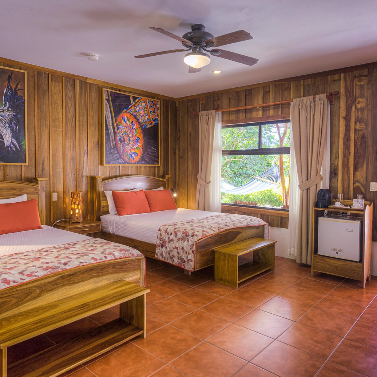 Hotel room with two beds, rooster paintings, wood paneling, and view of outdoor hammock.
