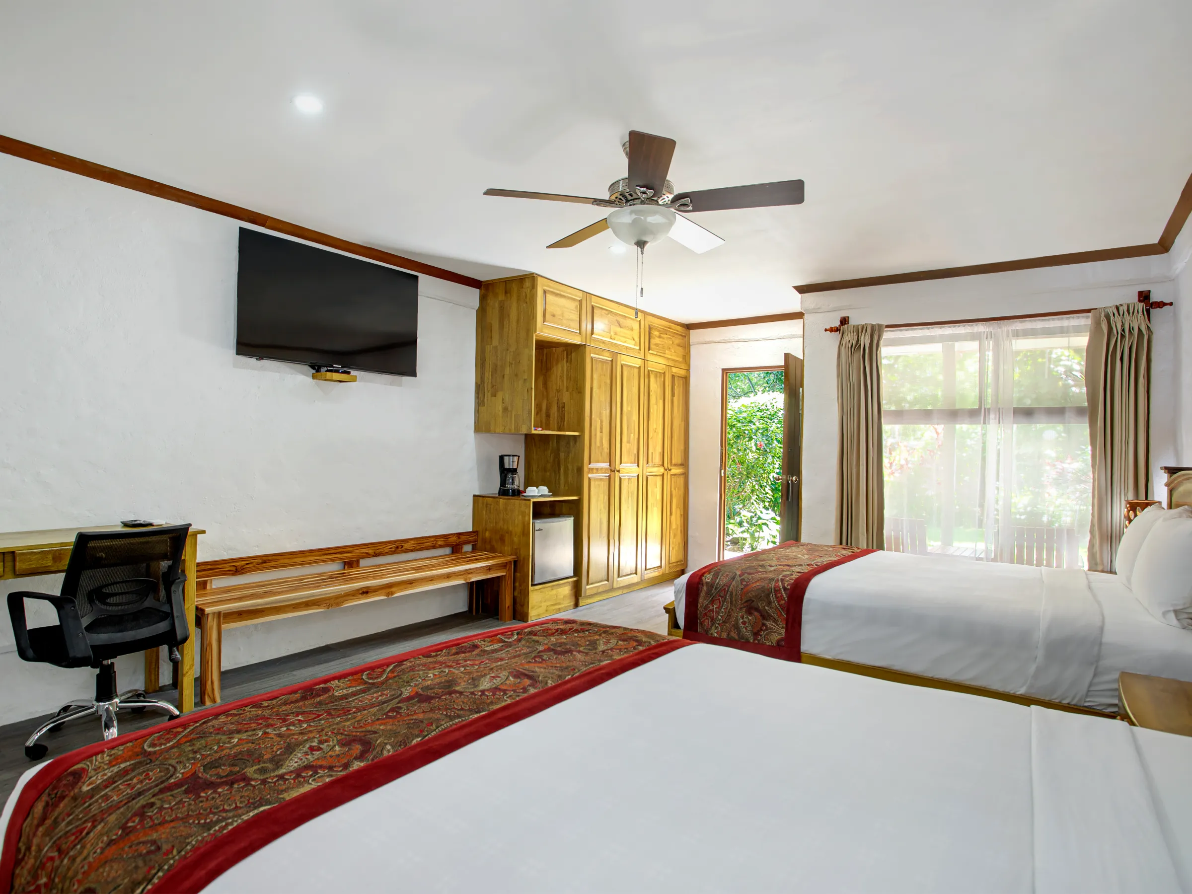 Hotel room with two beds, TV, desk, chair, ceiling fan, and large window with garden view.