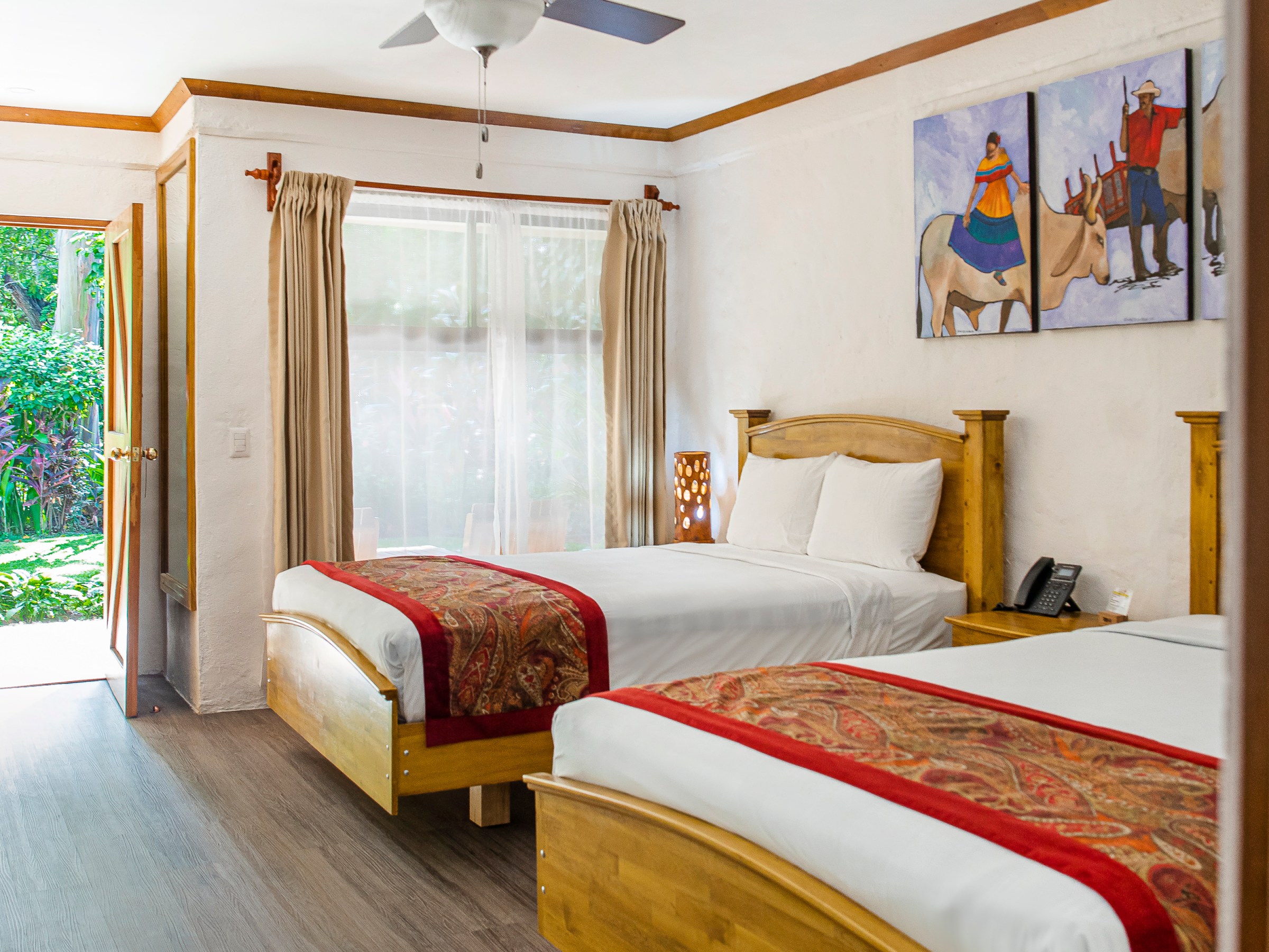 Room with two beds, ceiling fan, art on wall, and open door to garden.