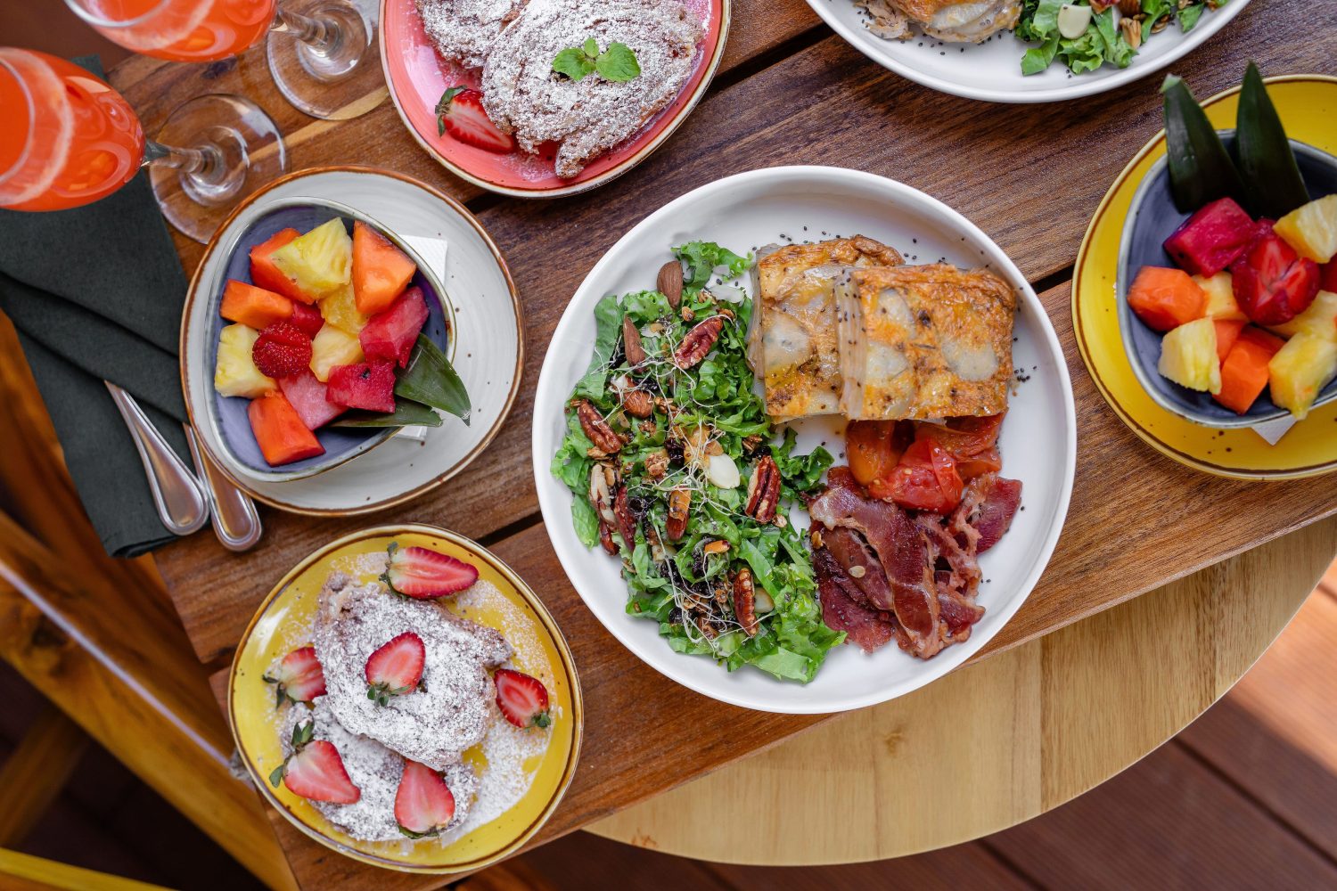 Brunch table with fruit bowls, salad, pastries, and meat on wooden surface.