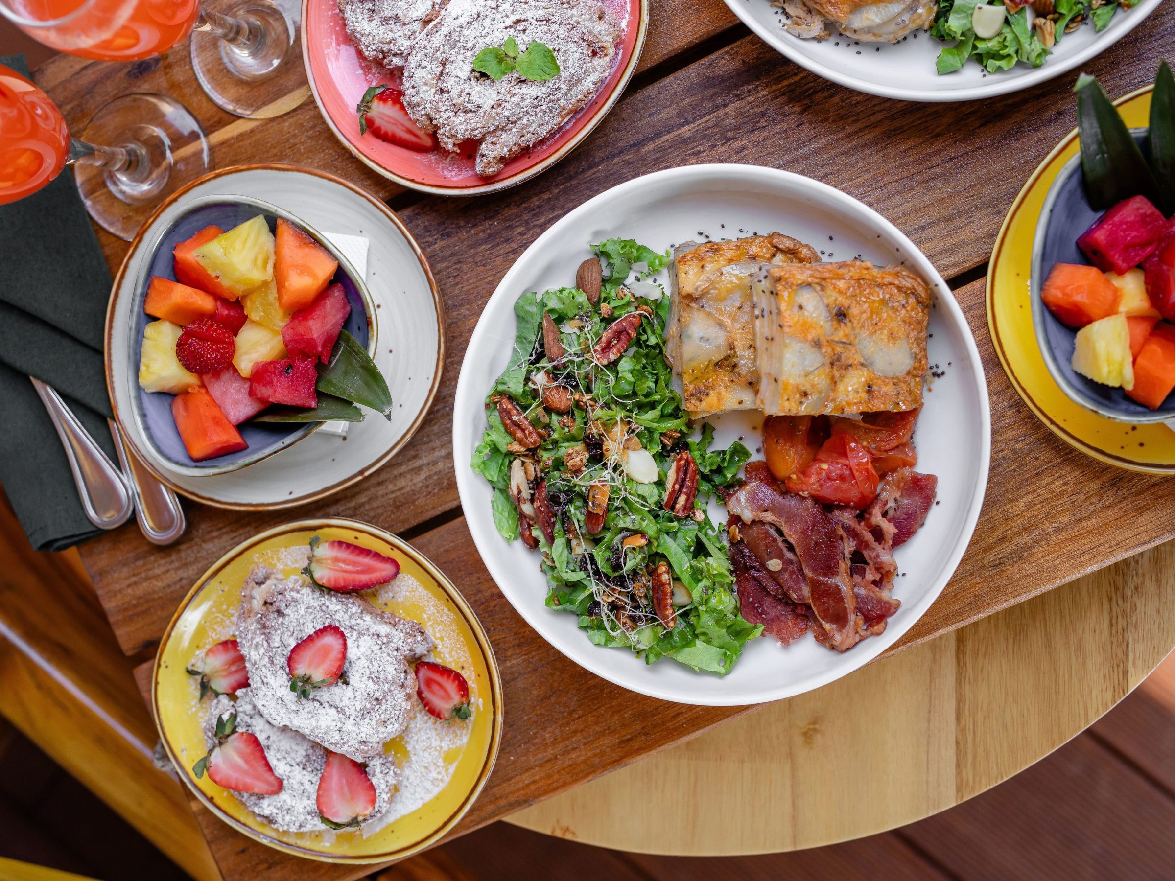 Brunch table with fruit bowls, salad, pastries, and meat on wooden surface.