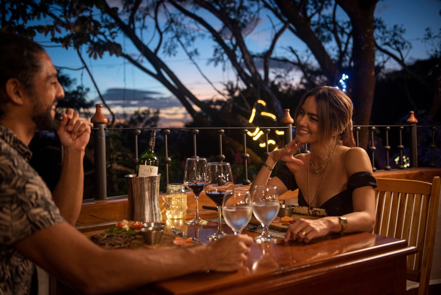 Couple dining outdoors at sunset, smiling with drinks on table.