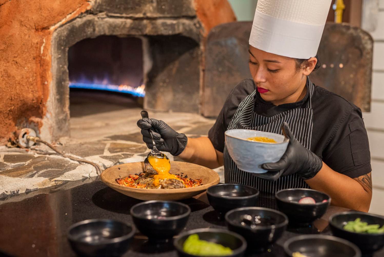 Chef in black gloves prepares dish in front of a wood-fired oven.