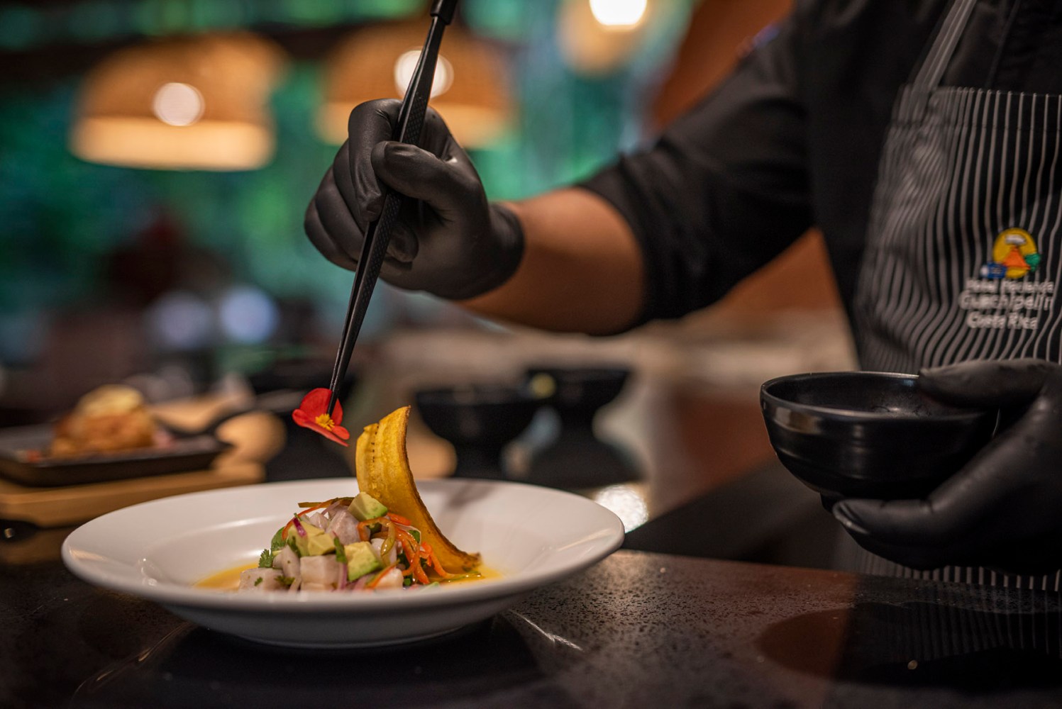 Chef in gloves garnishing a dish with tongs in a restaurant kitchen.