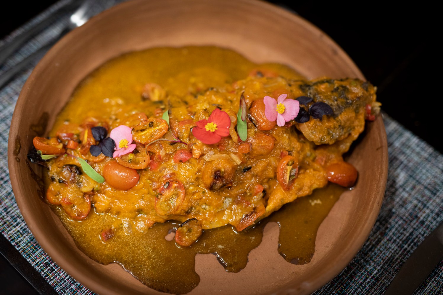 Colorful curry with vegetables and edible flowers in a brown dish on a textured placemat.