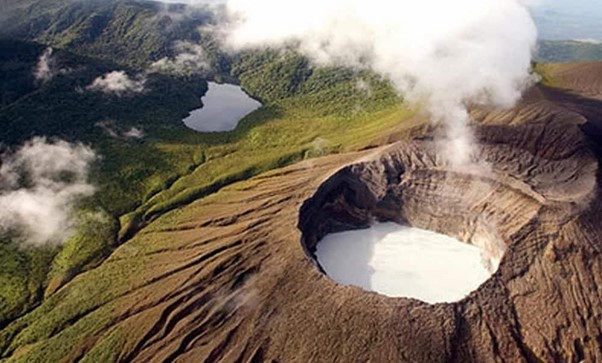 Aerial view of a volcanic crater with steam, surrounded by green forest.