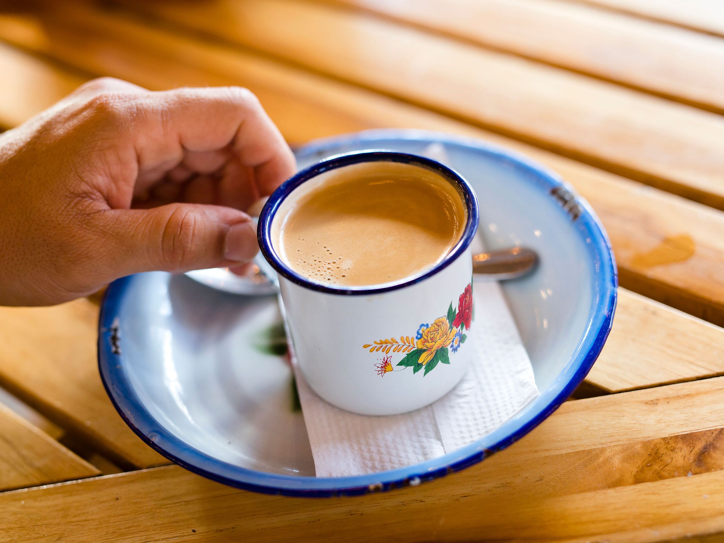 Hand holding floral mug of coffee on saucer with napkin.
