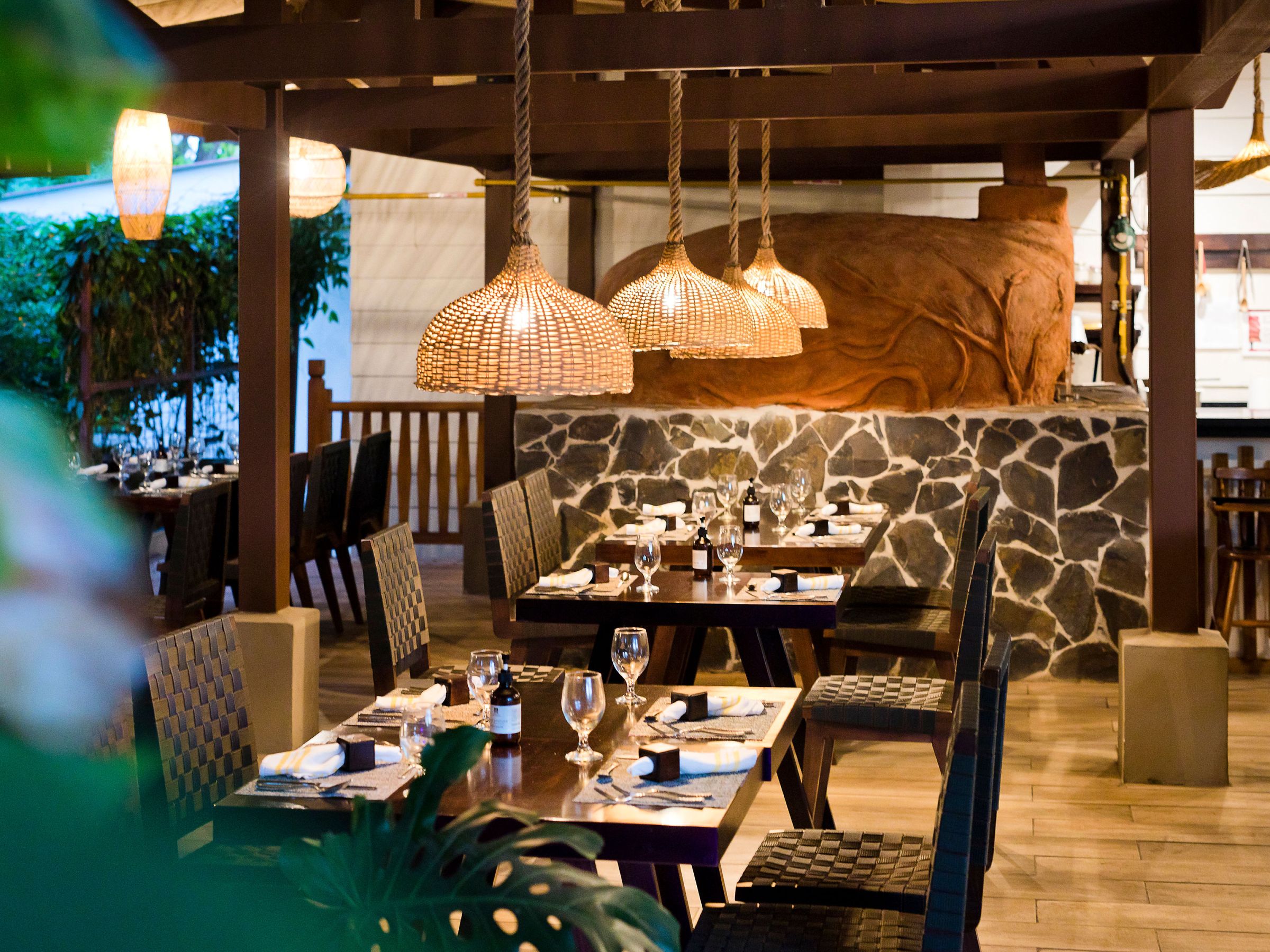 Empty restaurant with wooden tables, wicker lamps, and a stone wall.