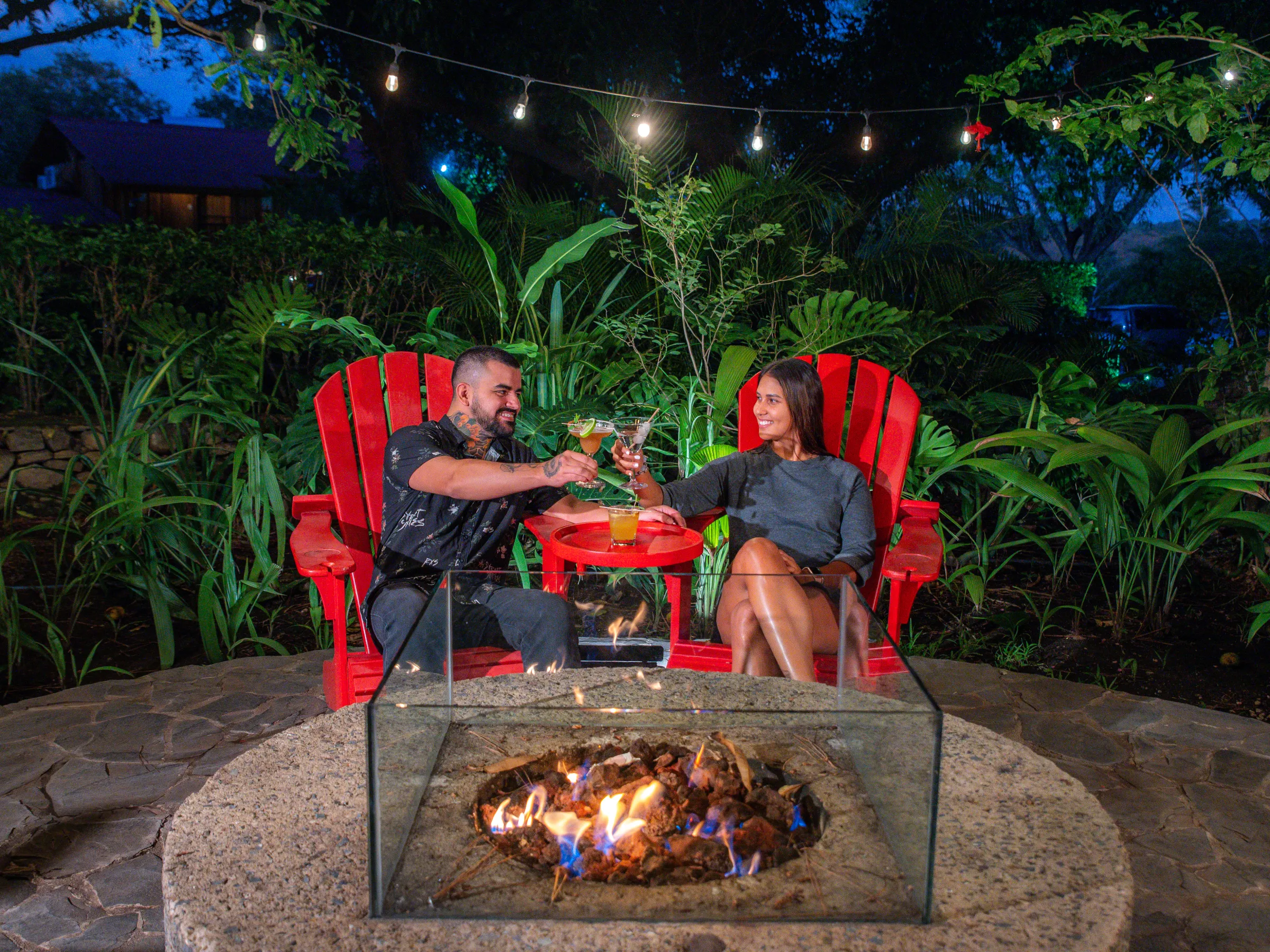 Couple toasting drinks by a fire pit with string lights and lush greenery around.