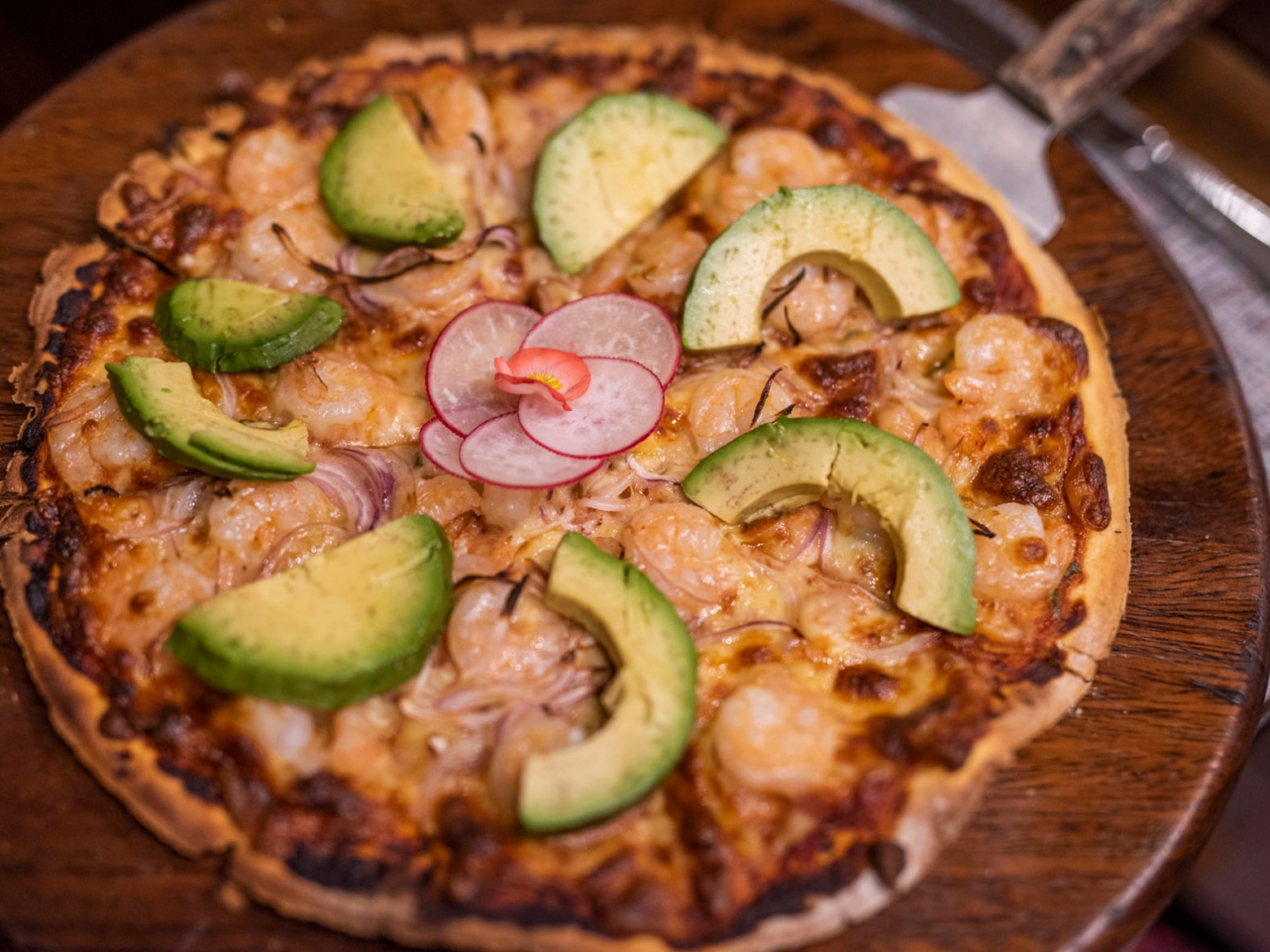 Pizza topped with avocado slices, shrimp, and radishes on a wooden tray.