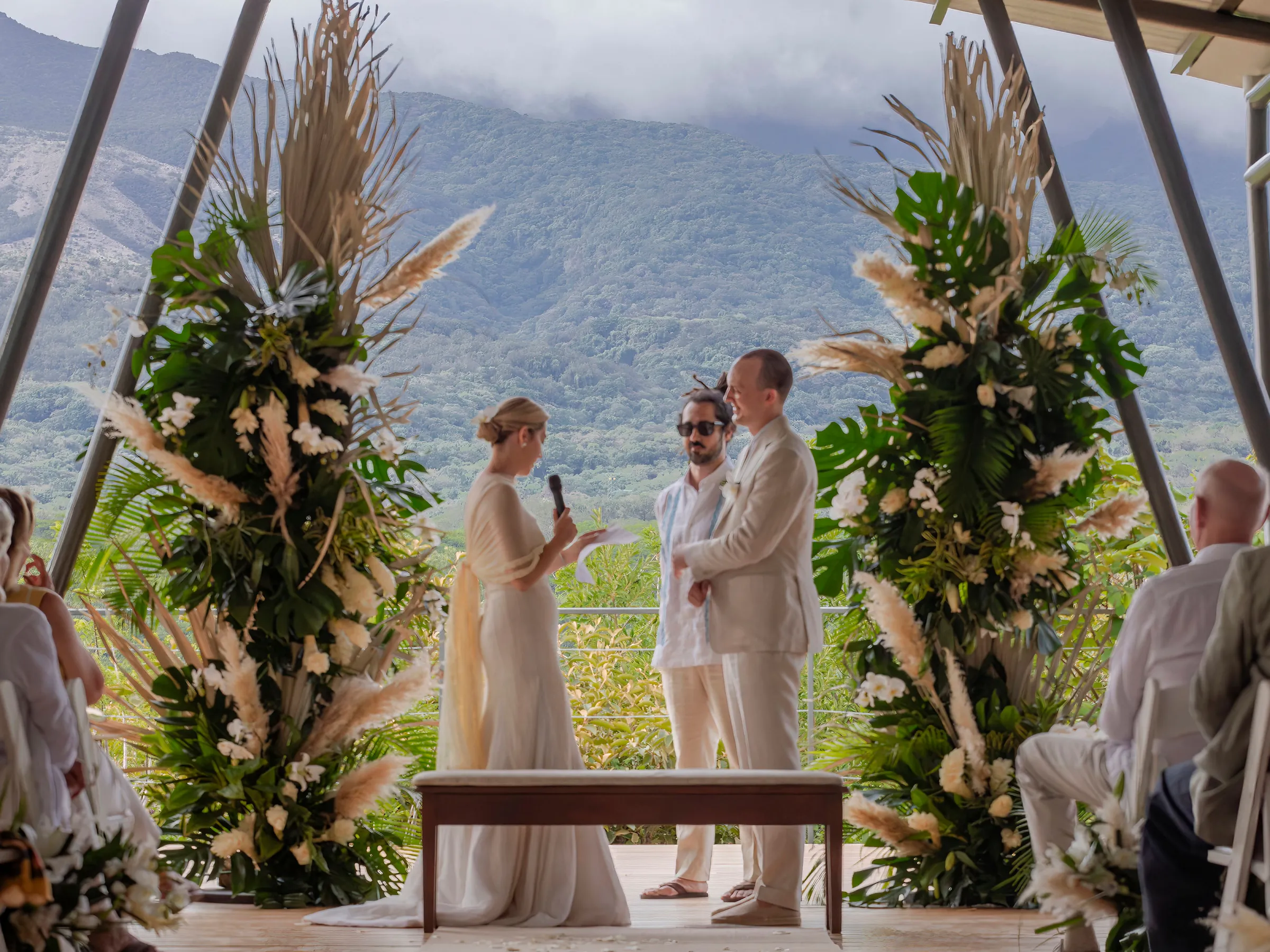 Outdoor wedding ceremony with couple and officiant, lush greenery backdrop.
