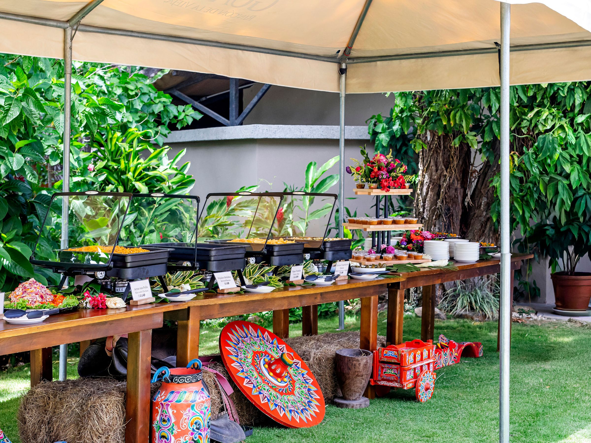 Outdoor buffet with various foods under a canopy, surrounded by greenery.