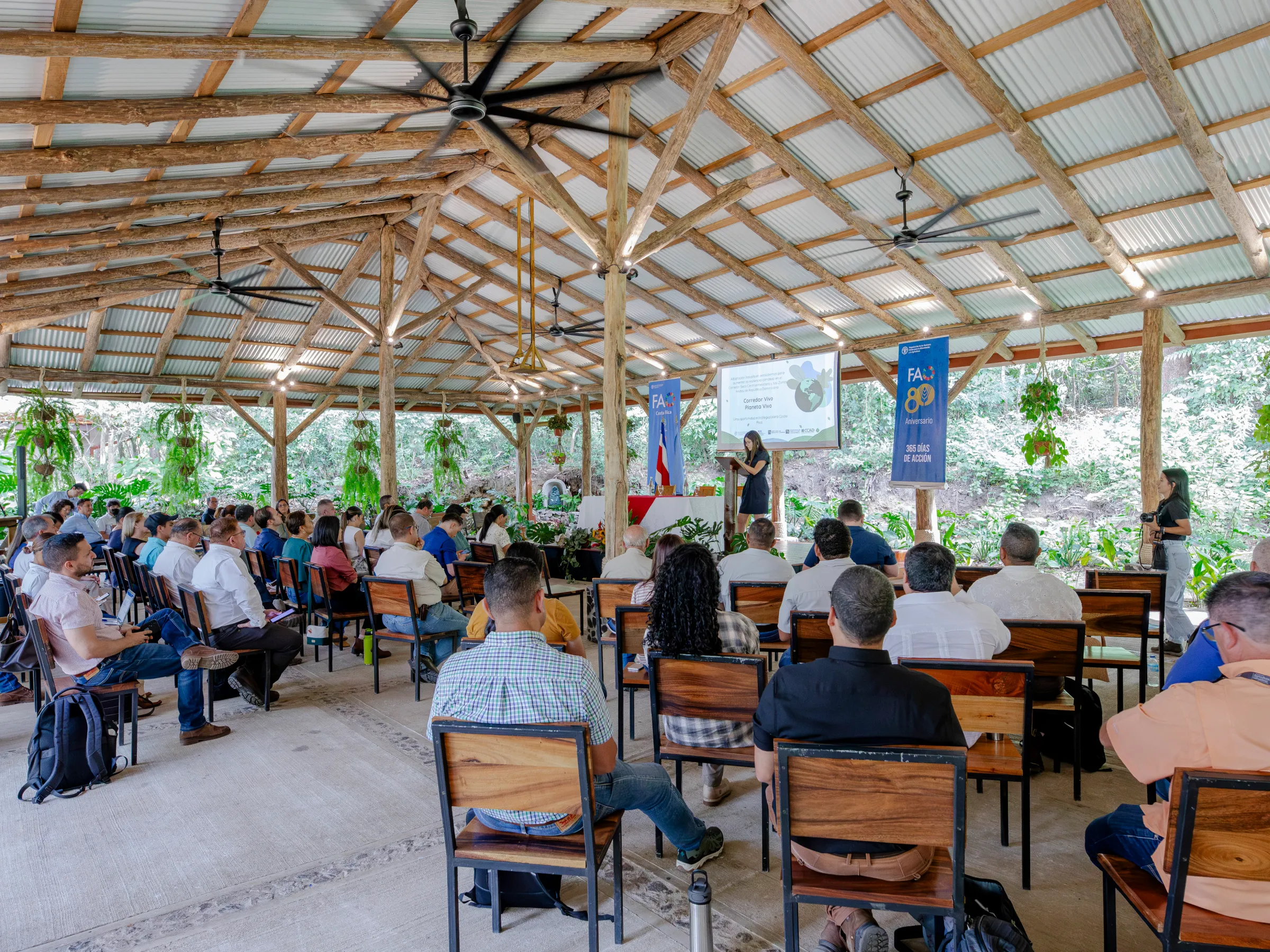 Conference in a rustic open-air setting, attendees seated, speakers at the front.