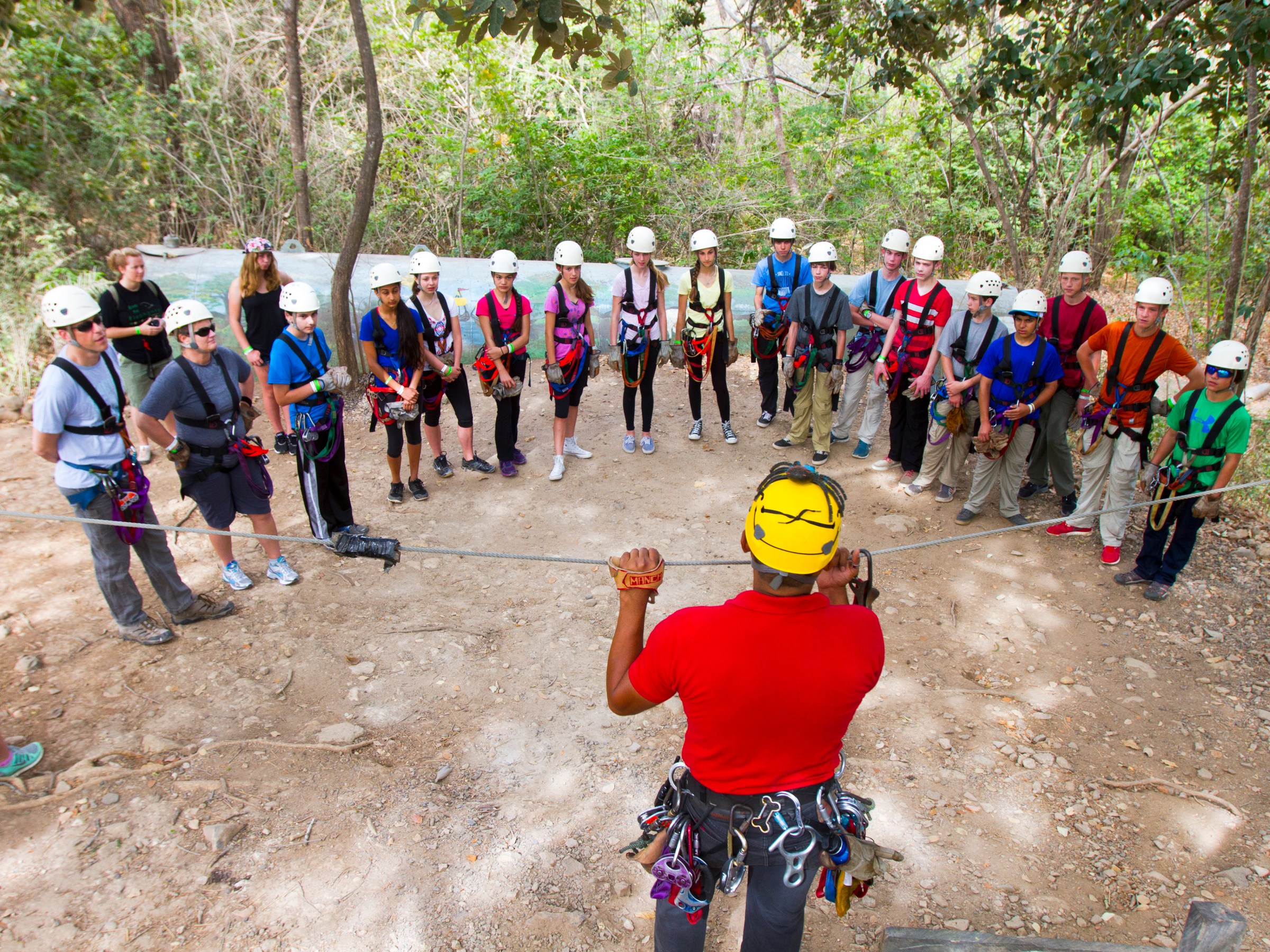 Instructor demonstrating zip line to a group wearing helmets and harnesses outdoors.