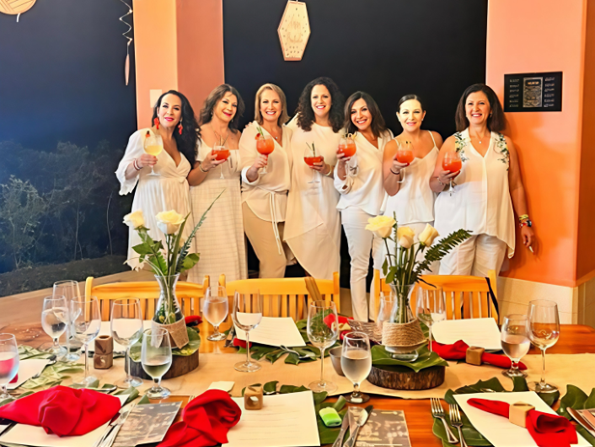 Eight people in white holding drinks, standing by a decorated table at a festive gathering.