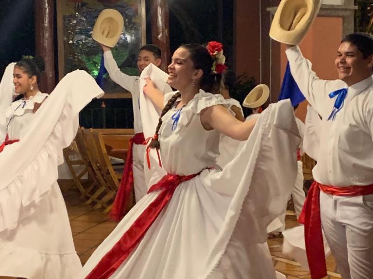Dancers in traditional white outfits with hats perform lively indoor dance.