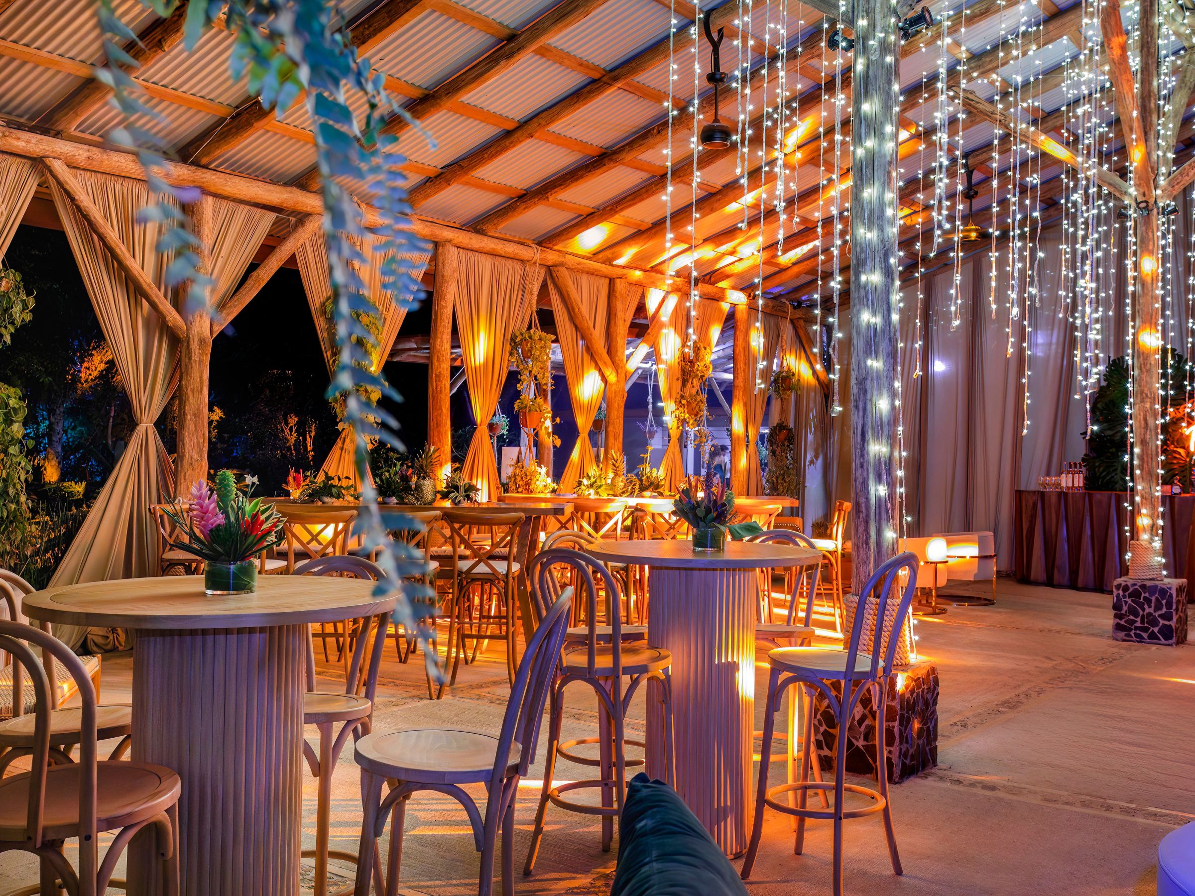 Stylish outdoor lounge with tables, hanging lights, and plants in warm lighting.