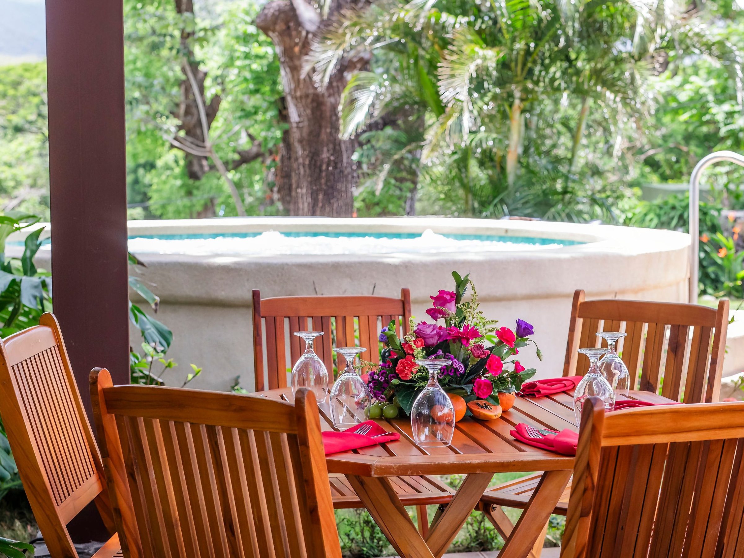 Outdoor wooden dining set with tableware, near a garden and circular spa.