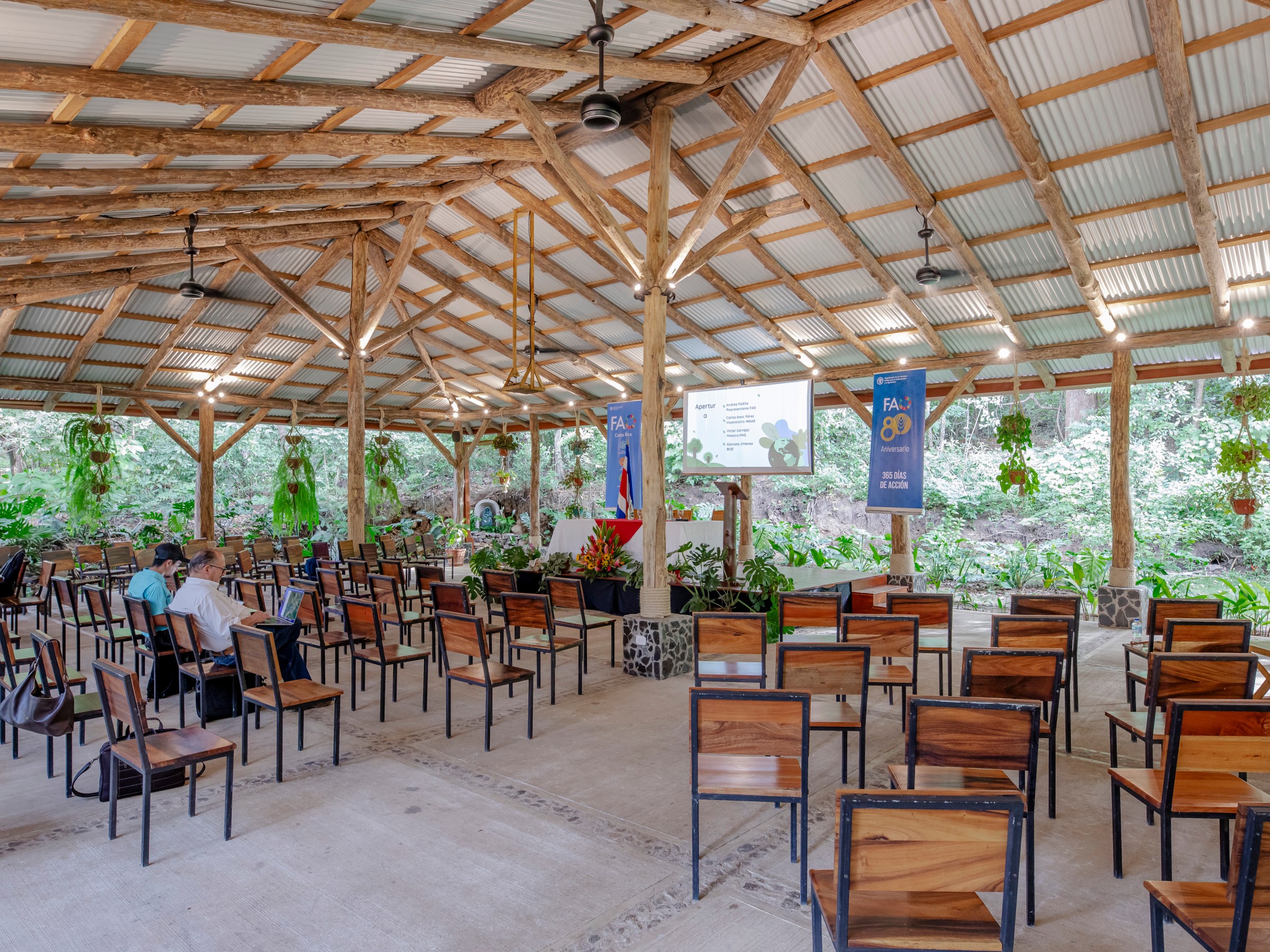 Outdoor event space with wooden chairs, screen, and tropical plants.