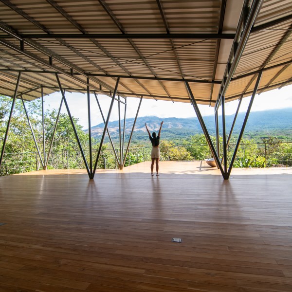 Person standing with raised arms on a wooden deck under a roof, overlooking mountains and greenery.