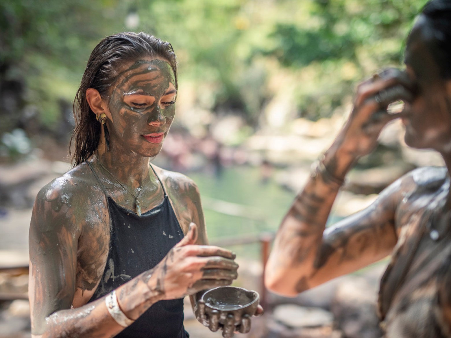 Two people covered in mud, outdoors near a water body, applying more mud to their skin.