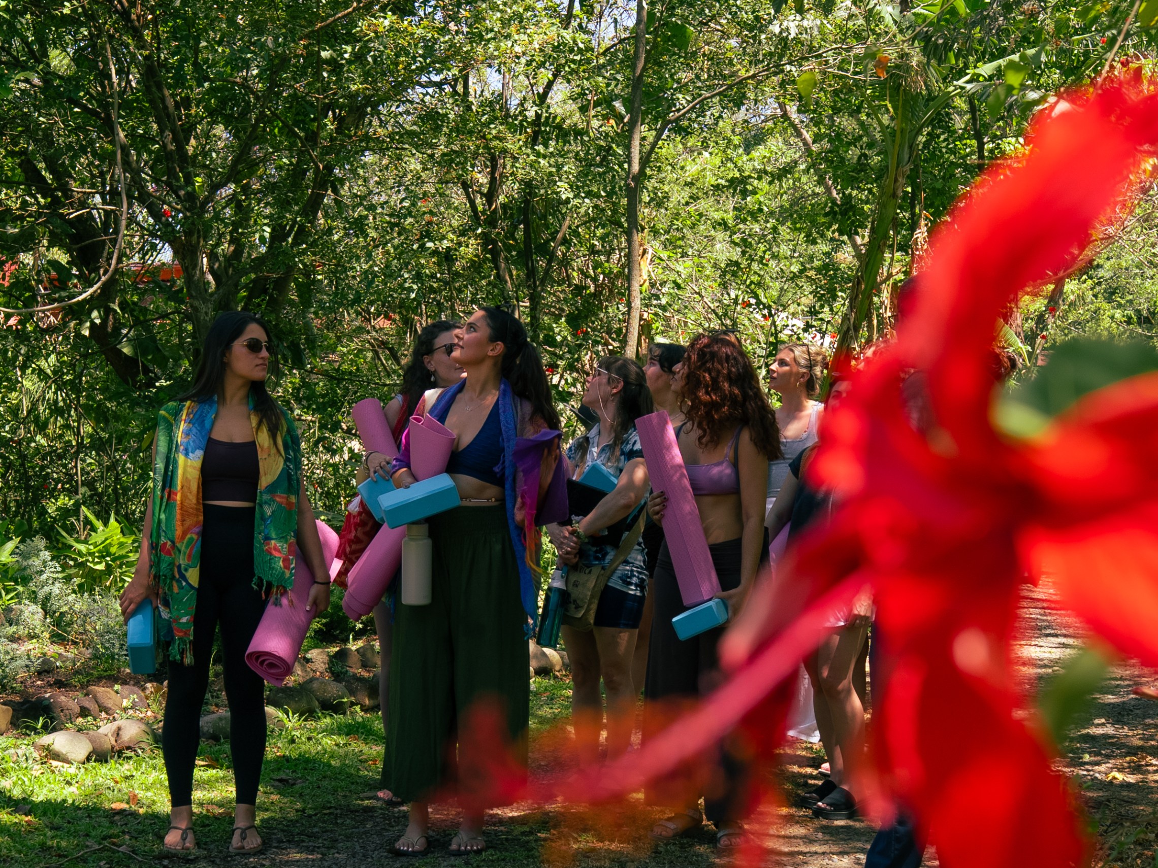 Group of people with yoga mats standing on a path surrounded by trees and flowers.