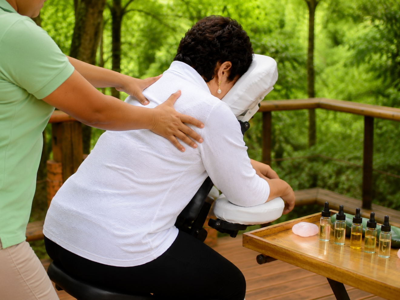 Person receiving outdoor massage, therapist standing, surrounded by trees and massage oils.