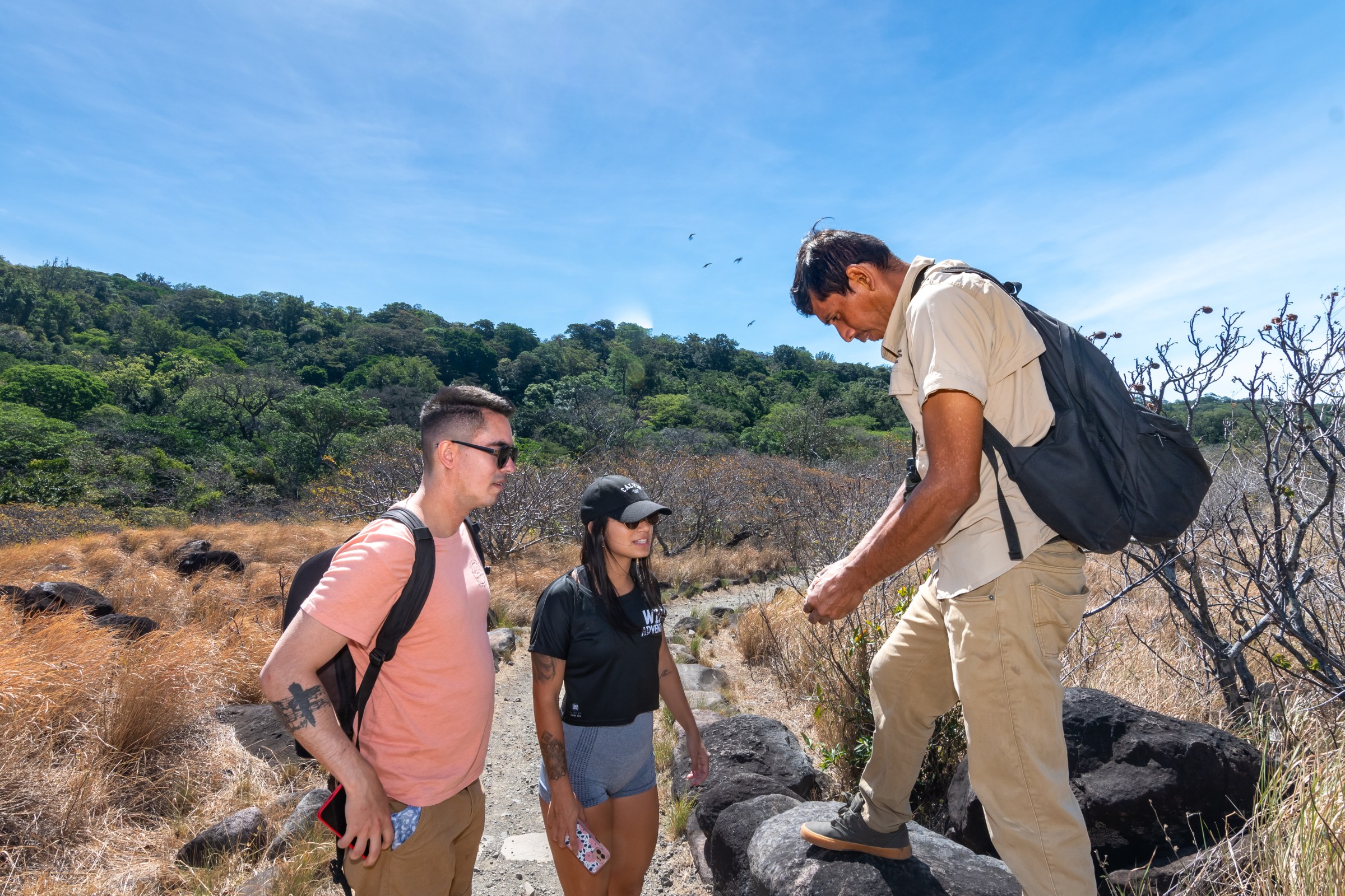 Three people hiking in a dry, rocky landscape with trees in the background under blue sky.